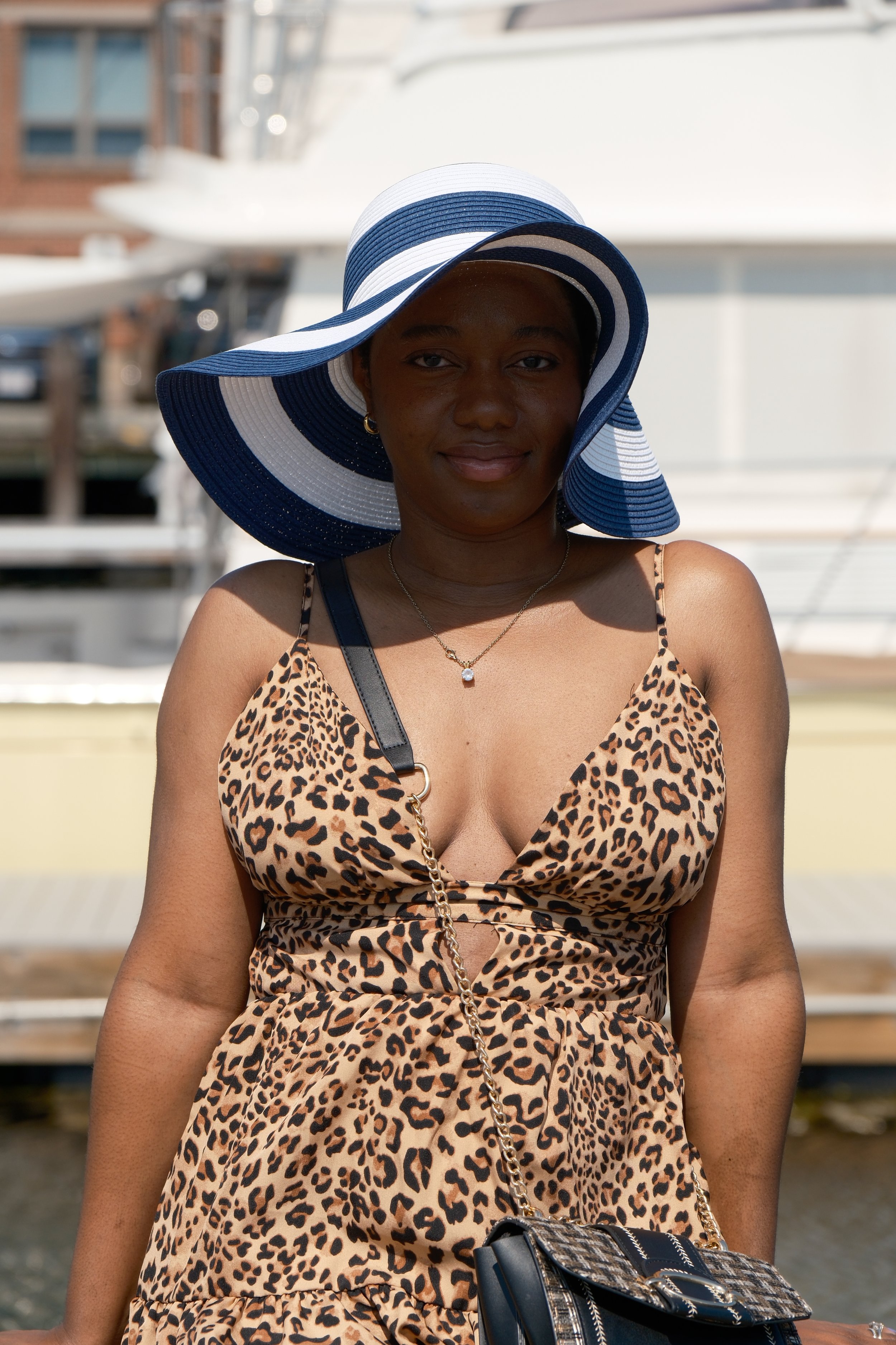 A woman wearing a large, wide-brimmed, blue and white striped sun hat, a leopard print dress, a gold necklace, and a black handbag with a chain strap, standing outdoors in front of a marina with boats.