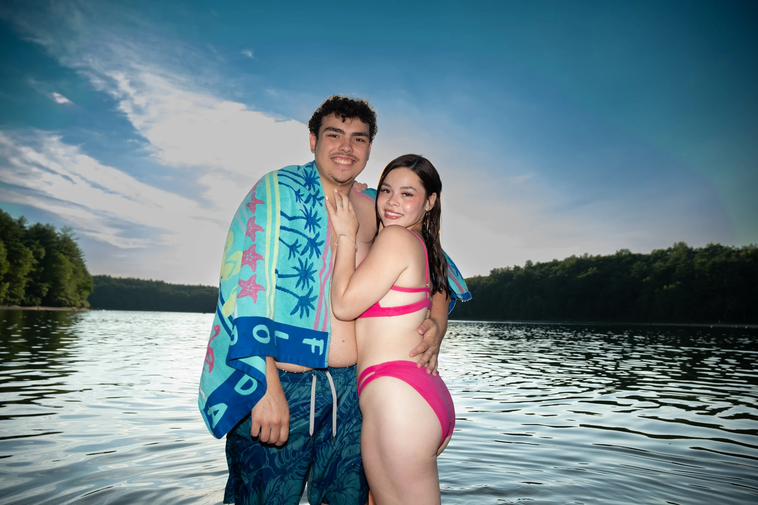 A smiling young man and woman stand close together near a lake, with the man wrapped in a colorful towel and the woman in a pink swimwear. Background shows a calm lake with trees and a partly cloudy sky.