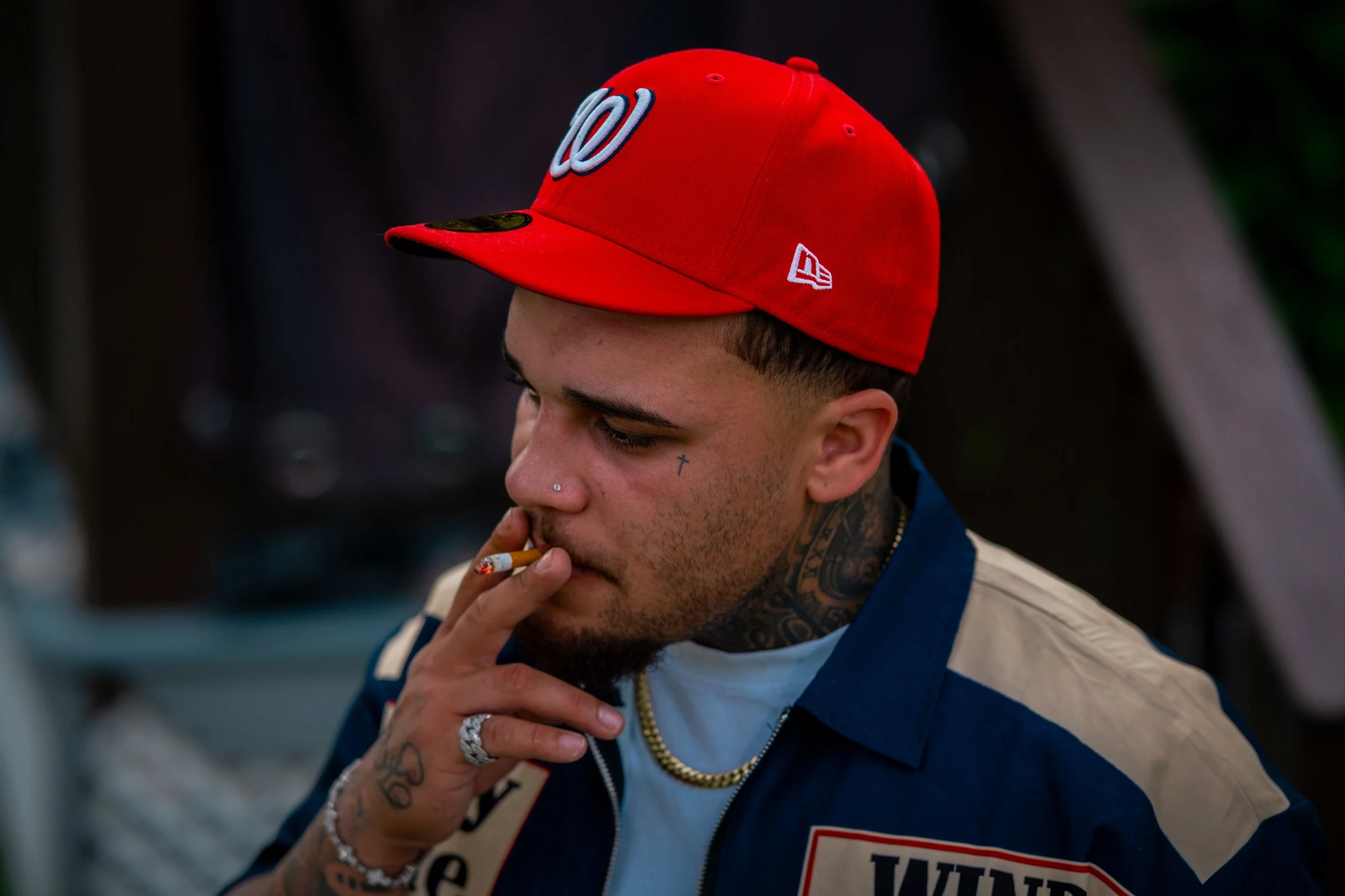 A young man with tattoos, piercings, and a nose ring wearing a red baseball cap with a Washington Nationals logo, smoking a cigarette, and wearing a blue and beige jacket.