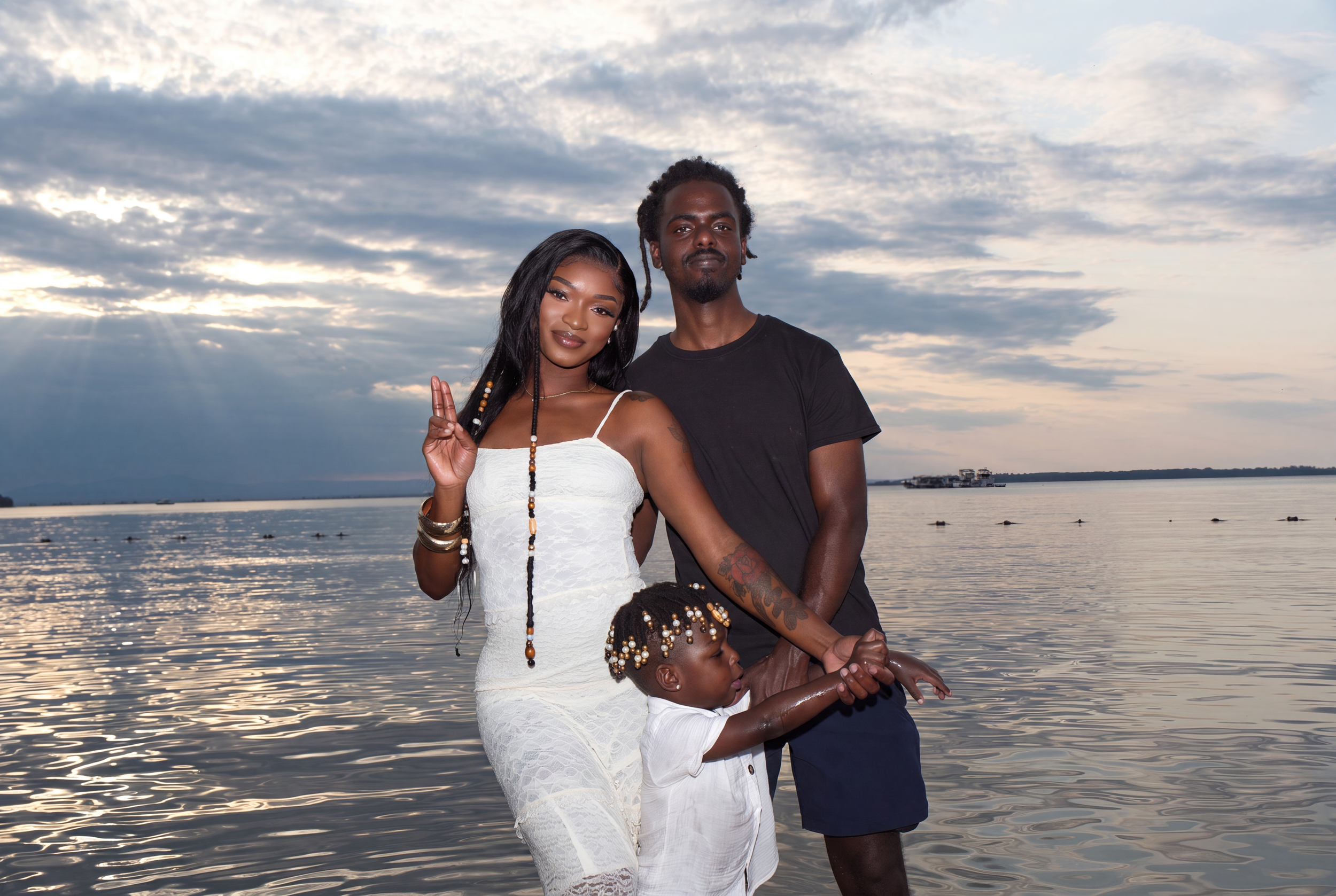 A family of three standing in front of a body of water with a cloudy sky, some of the family members are making peace signs.