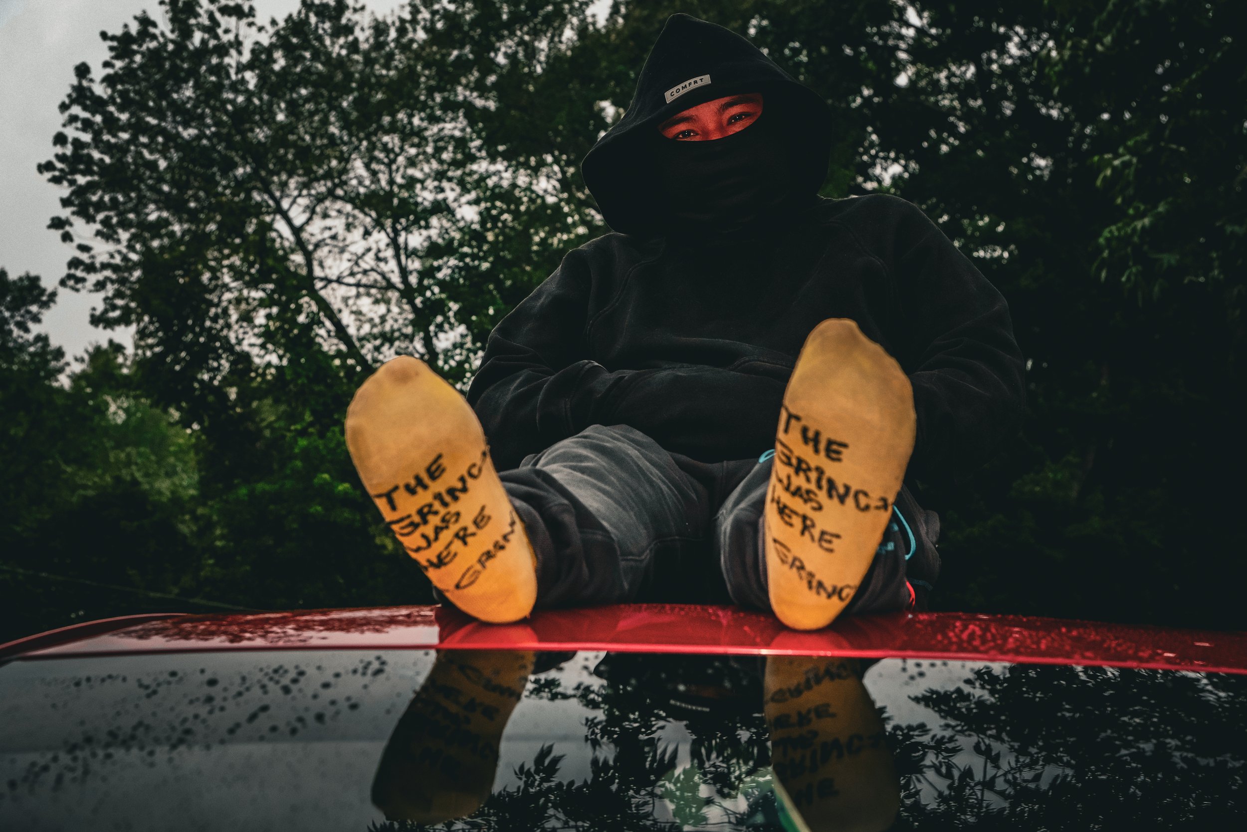Person sitting outdoors on a red surface with trees in the background, wearing a black mask and hoodie, with their feet facing the camera. The soles of the shoes have handwritten messages that read 'THE STRING WAS HERE' and other words.