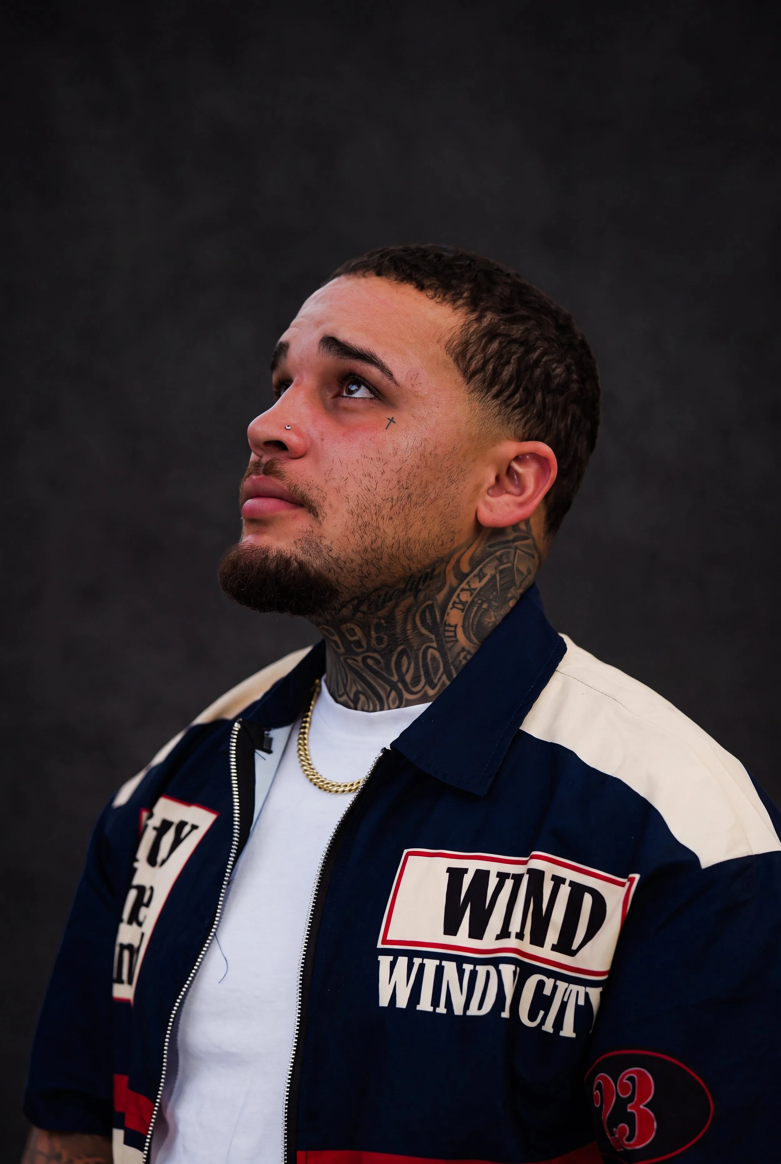 A young man with short dark hair and tattoos on his neck and face looking upward. He has a nose piercing and wears a gold chain. He is wearing a jacket with patches that say 'WINDY CITY' and '23', over a white T-shirt, against a dark background.