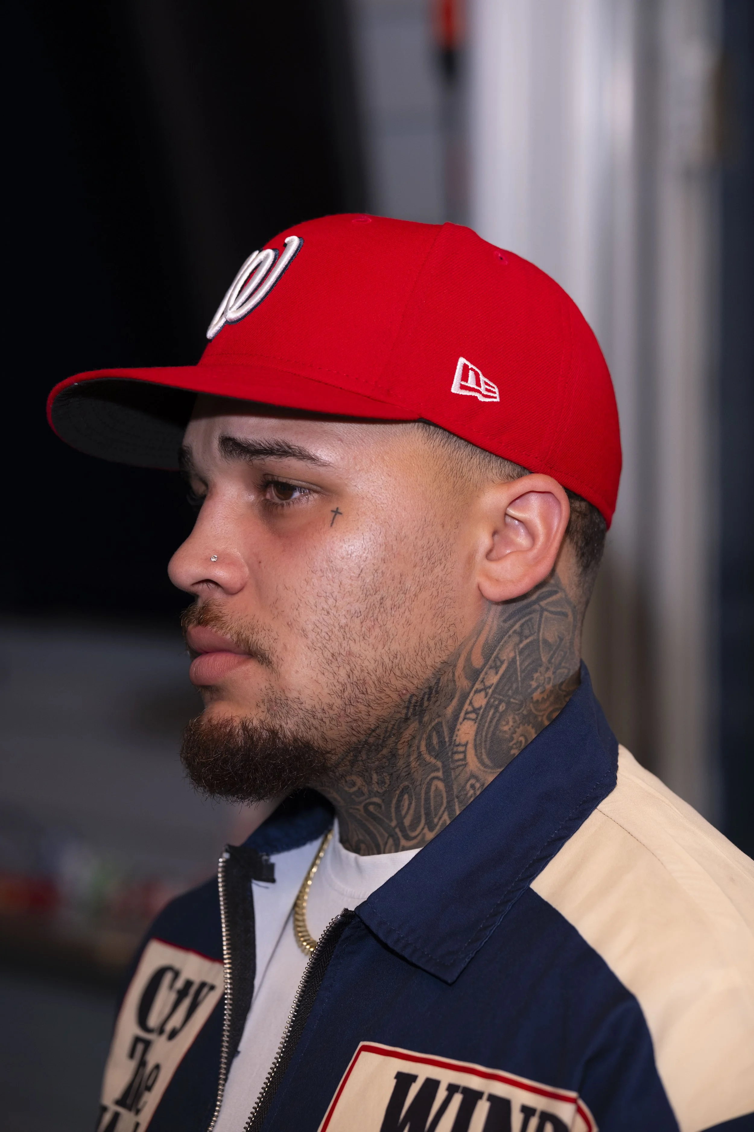 Close-up profile of a young man with tattoos on his neck, wearing a red baseball cap and a jacket with patches. The background is blurred.