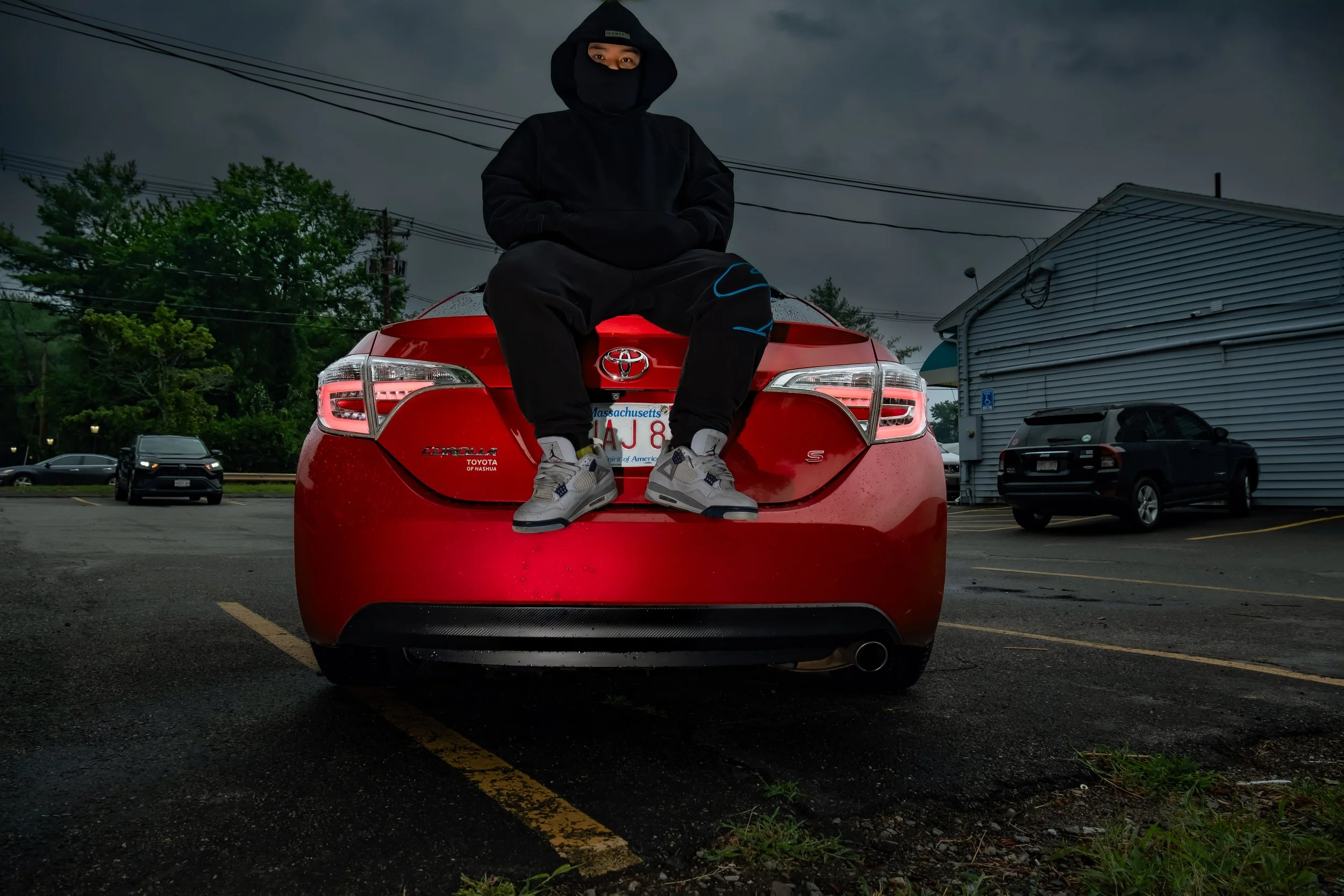 Person dressed in black sitting on the trunk of a red Toyota car in a parking lot on a cloudy day.
