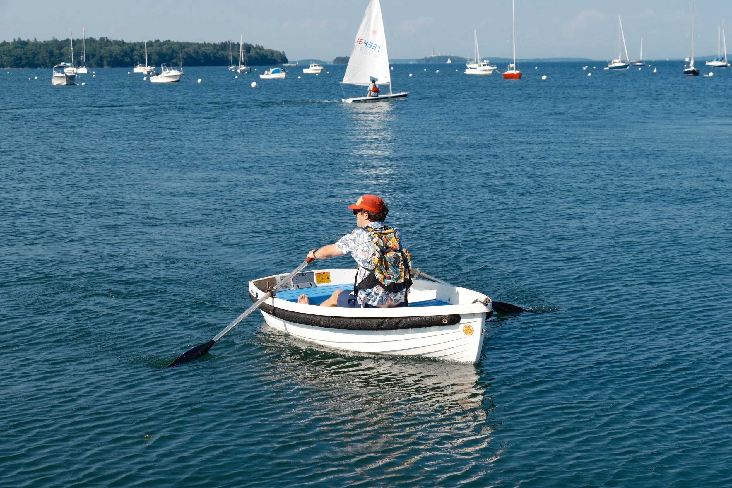 A person wearing a red cap and sunglasses sitting in a small white rowboat with an oar, floating on a calm body of water with many sailboats and yachts anchored in the distance, and a tree-covered coastline in the background.