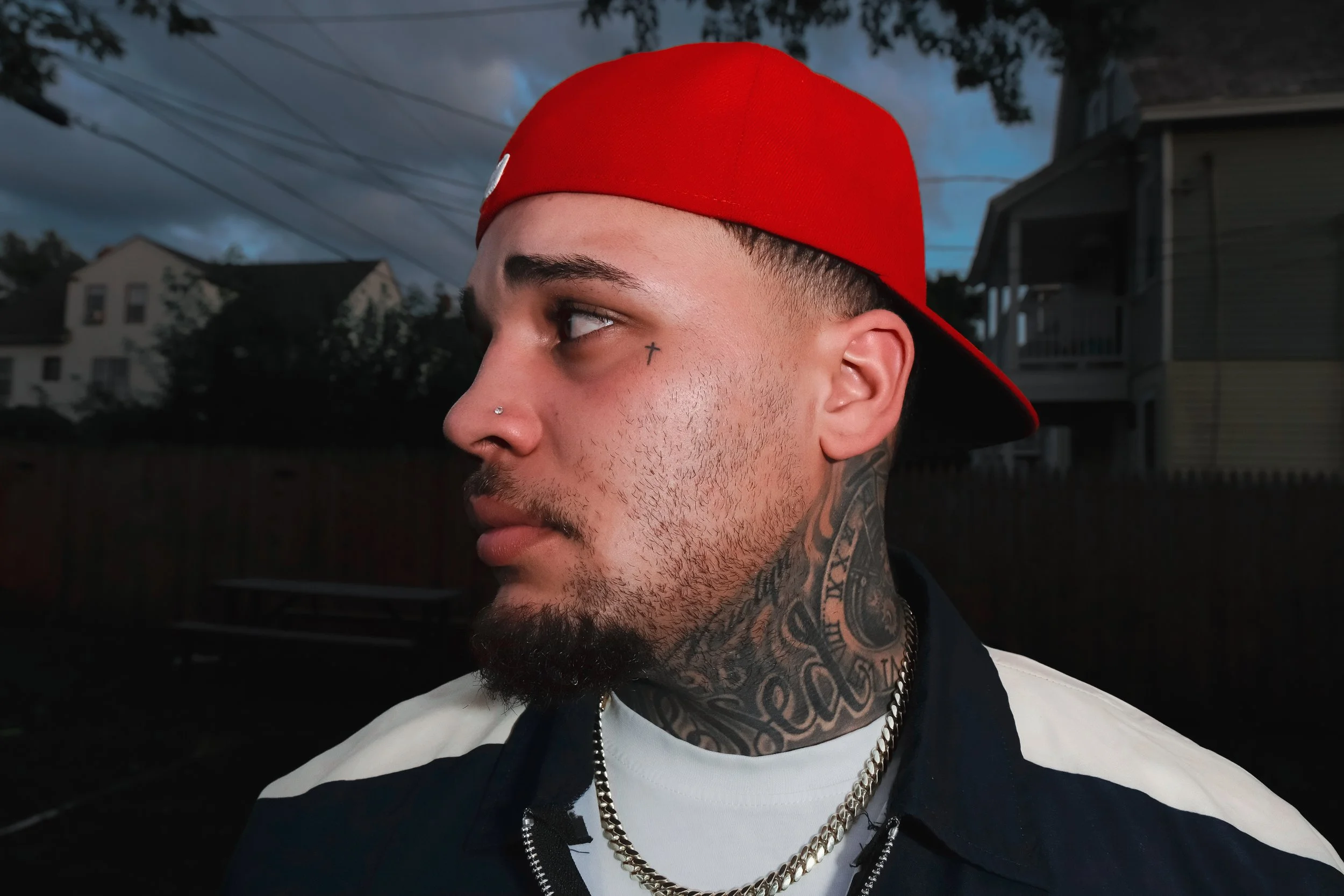 A young man with tattoos and facial piercings wearing a red cap, looking to the side with a background of houses and cloudy sky.