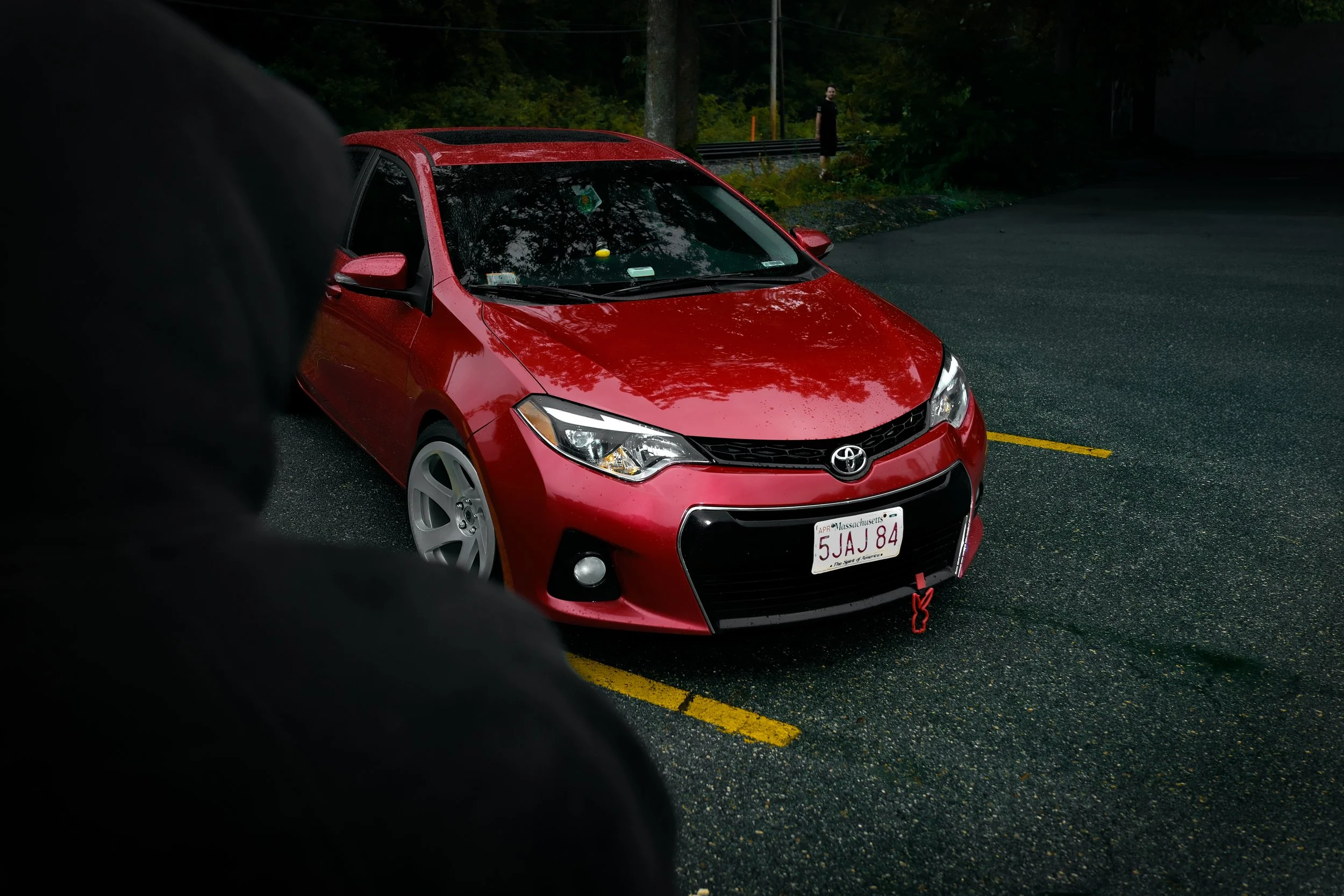 A red Toyota car parked on a wet road with a person standing in the background near a guardrail and trees.