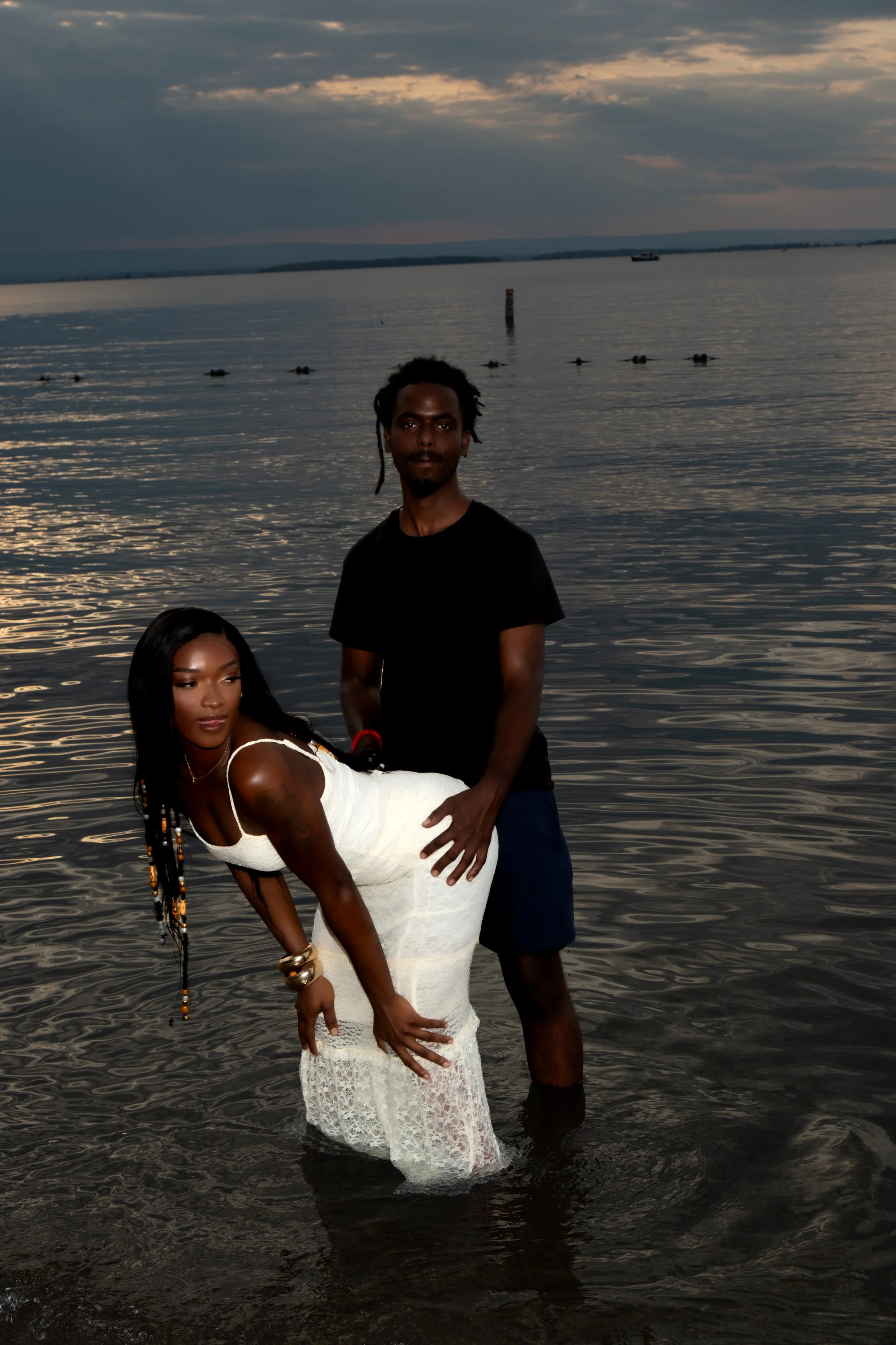 A woman in a white lace dress bends forward in the water, with a man standing behind her, holding her waist, by a lakeside at sunset. The sky is cloudy with a quiet lake and distant hills in the background.