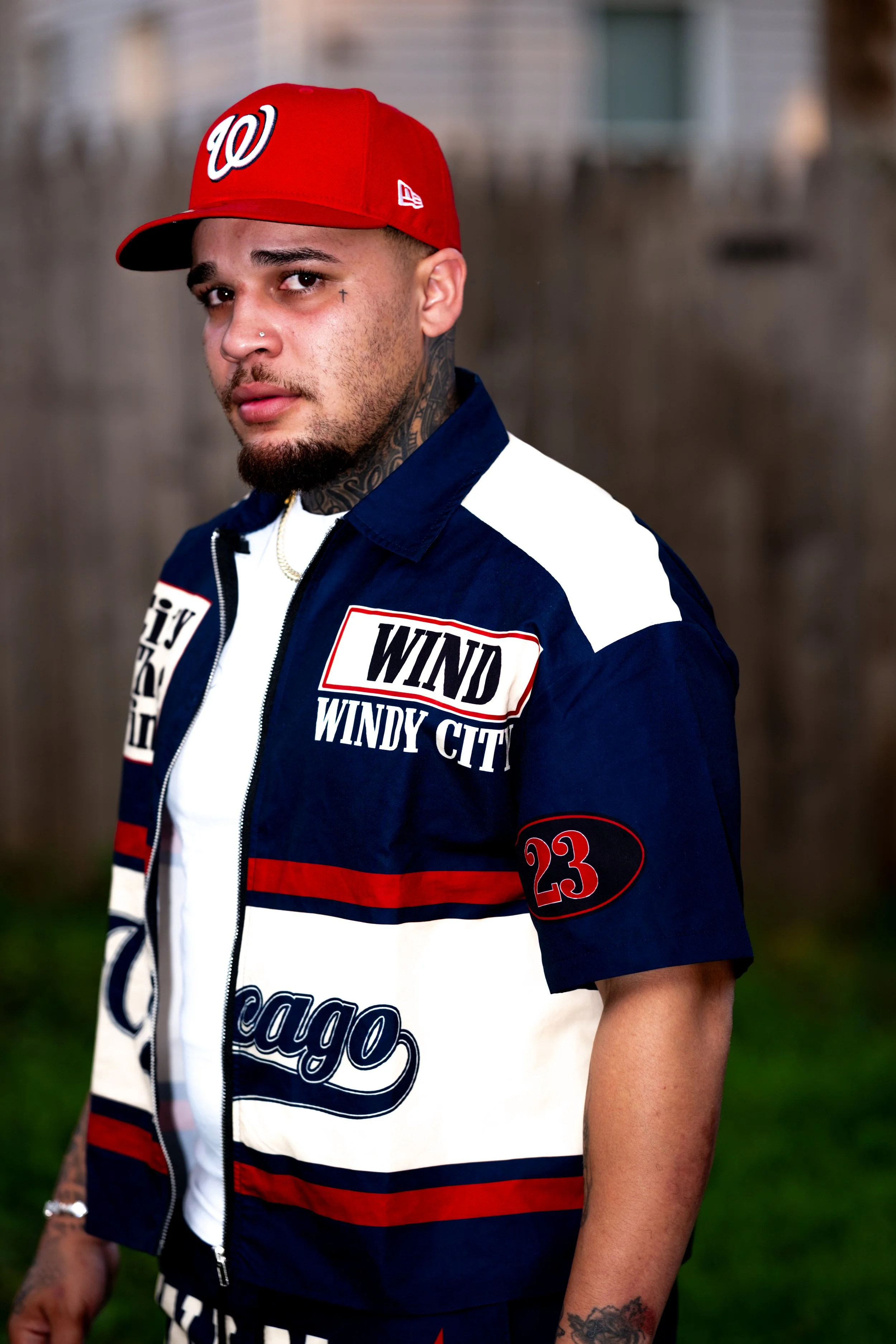 A young man with tattoos and facial jewelry wearing a red cap and a Chicago Bears-themed jacket with patches that say 'WINDY CITY' and '23', standing outdoors with a blurred background.