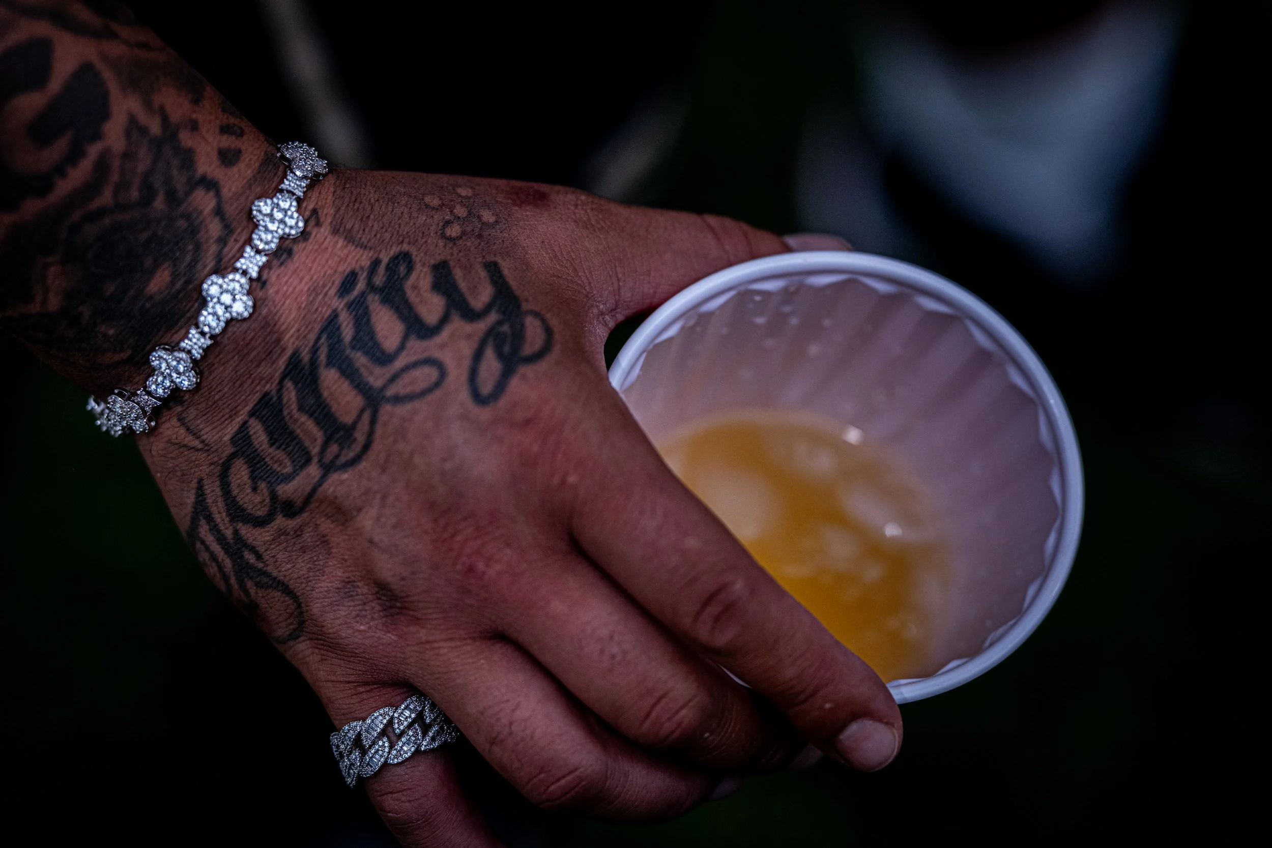 A person with tattooed hand wearing a diamond-studded bracelet and ring, holding a paper cup with a yellow beverage, against a dark background.