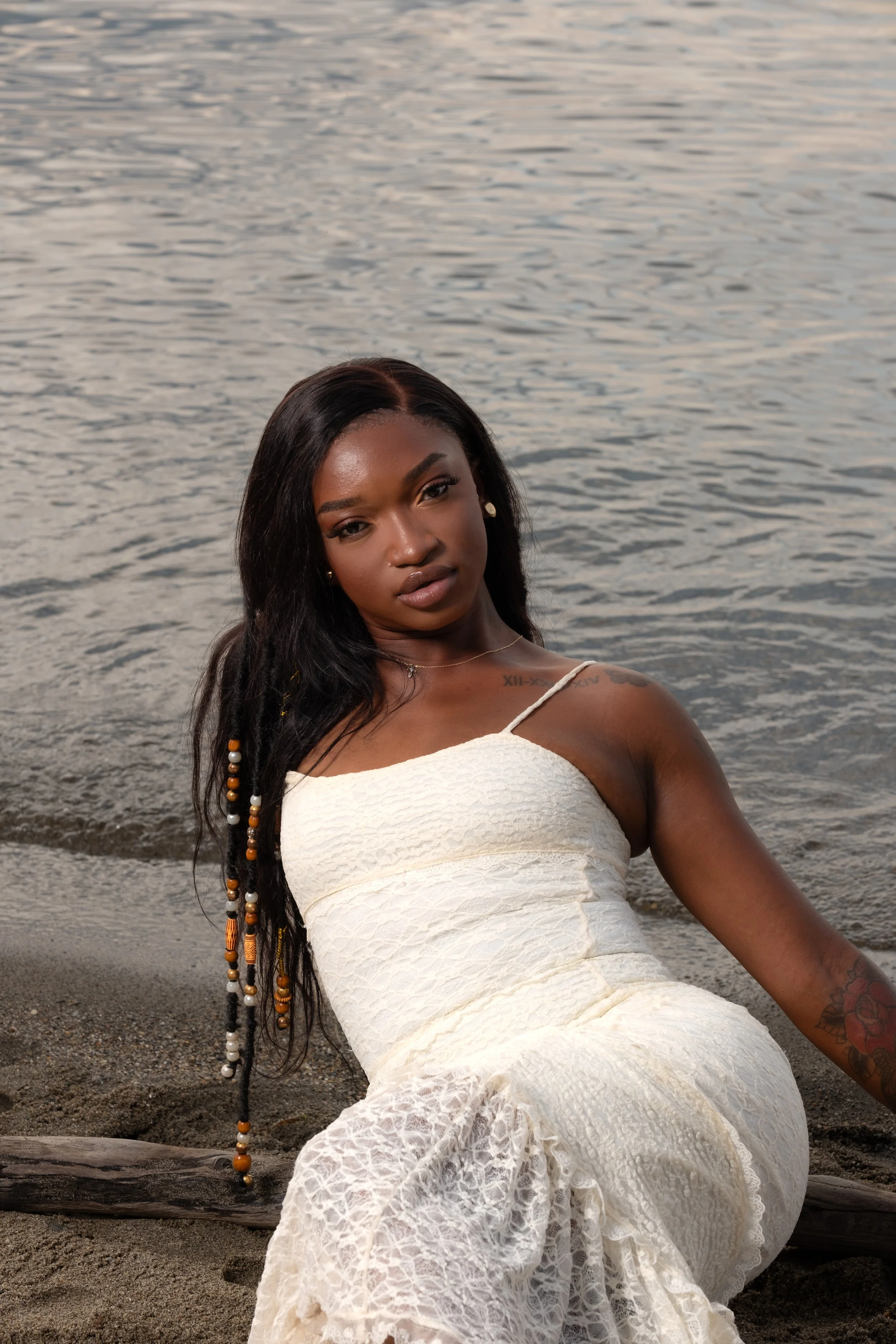 A woman sitting on beach sand near water, wearing a white lace dress with long black hair and beads, posing for a photo.