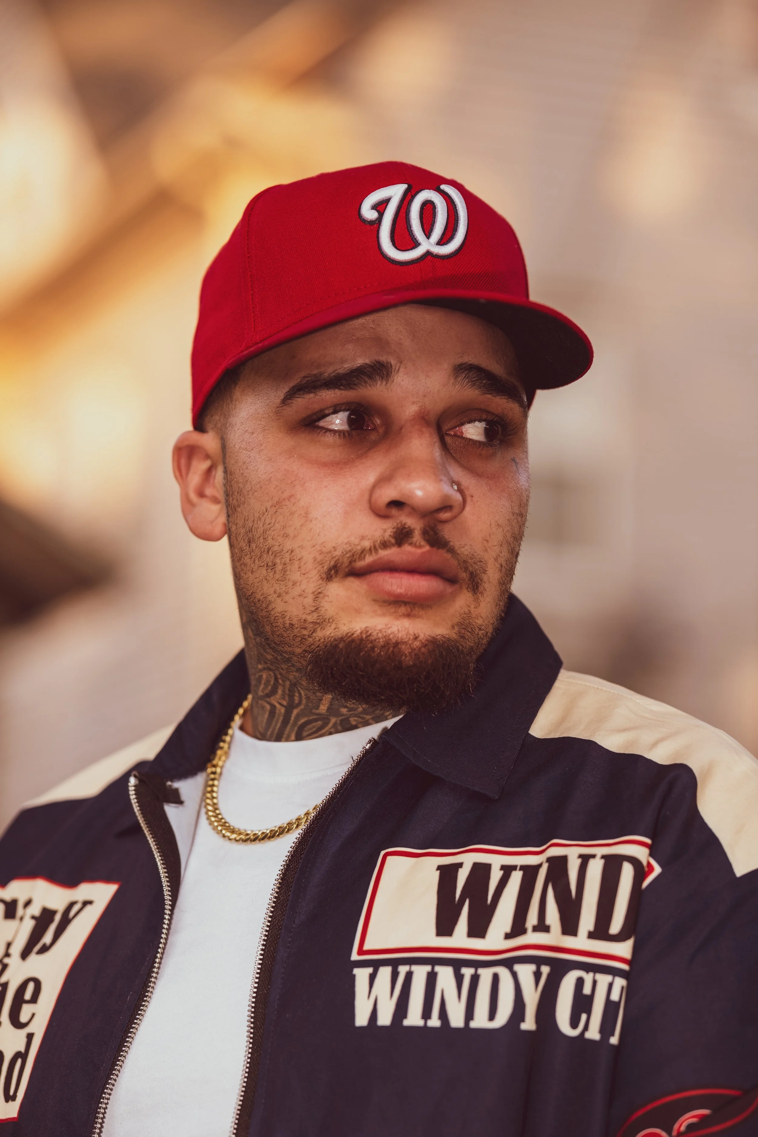 Young man with a beard and mustache wearing a red baseball cap with a white and black logo, a tattooed neck, gold chain, and a black and white jacket with a Windy City patch.