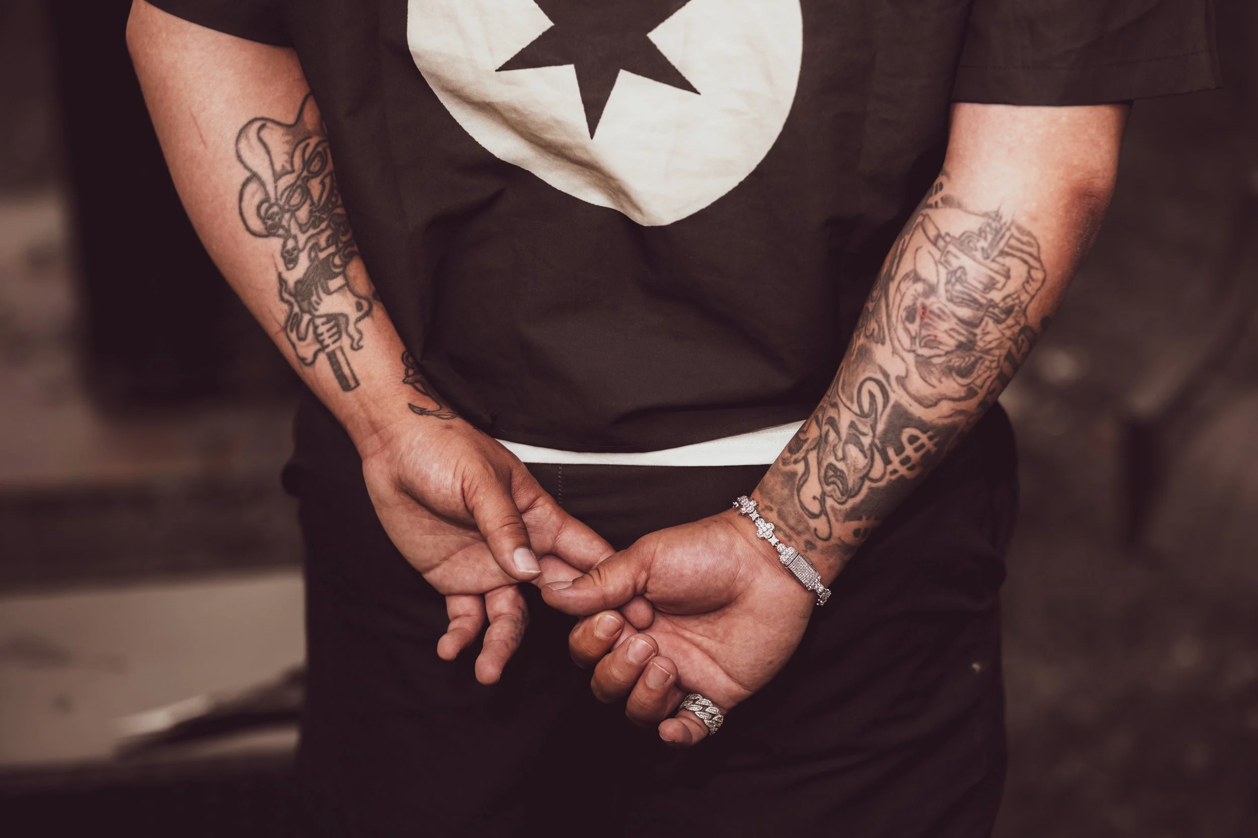 Close-up of a person with tattoos on both arms, wearing a black T-shirt with a white star within a circle design, and holding their hands together in front of them.