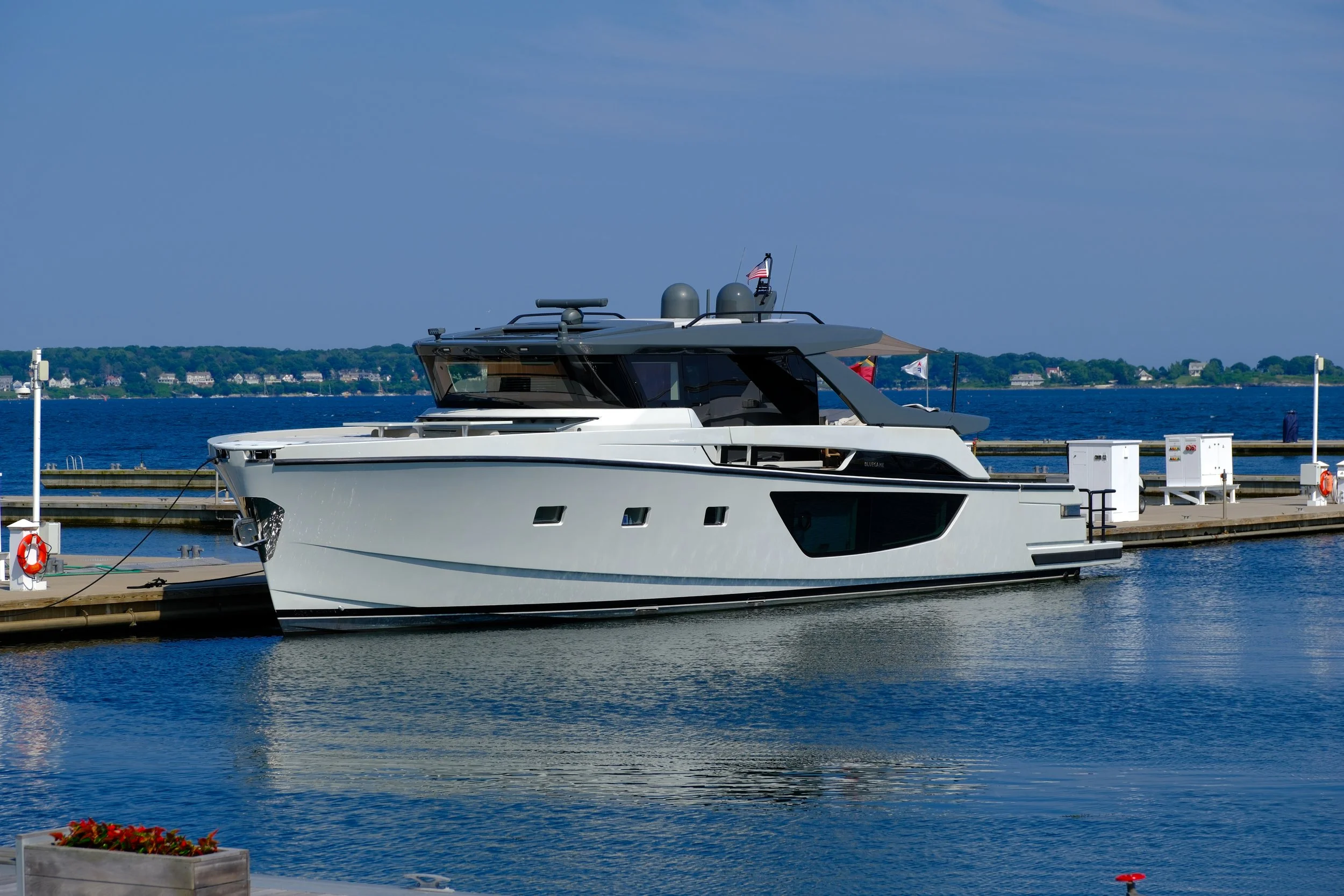 A luxury yacht docked at a marina on a clear day with calm waters and a shoreline with houses in the background.