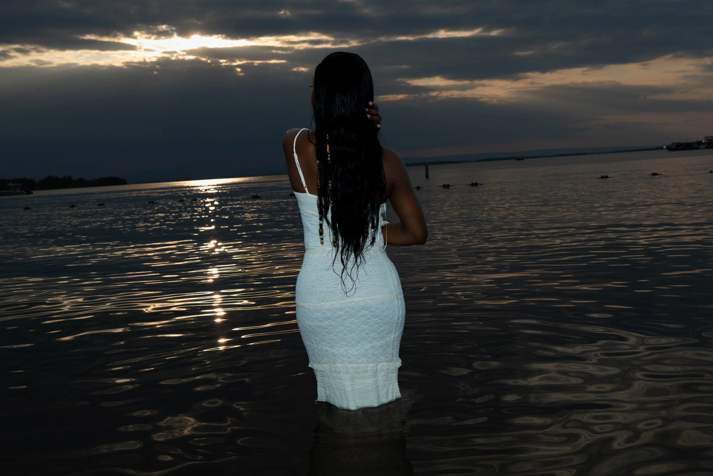 A woman with long black hair, wearing a white dress, stands in a body of water during sunset or sunrise, facing away from the camera, with clouds in the sky reflecting on the water surface.