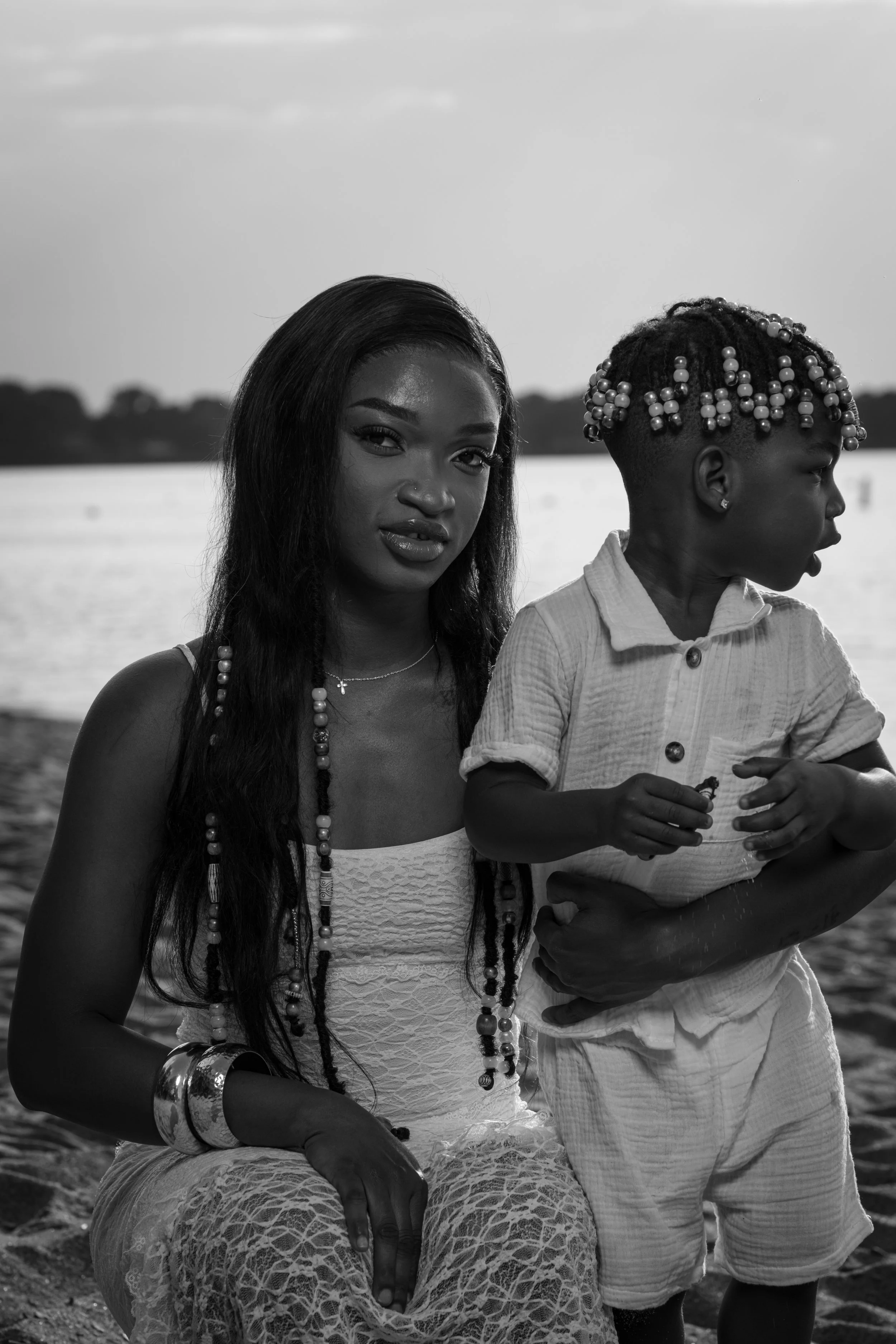 A black and white photo of a woman with long hair and accessories sitting by the water, with a young girl with beaded hair standing beside her, both looking in different directions.