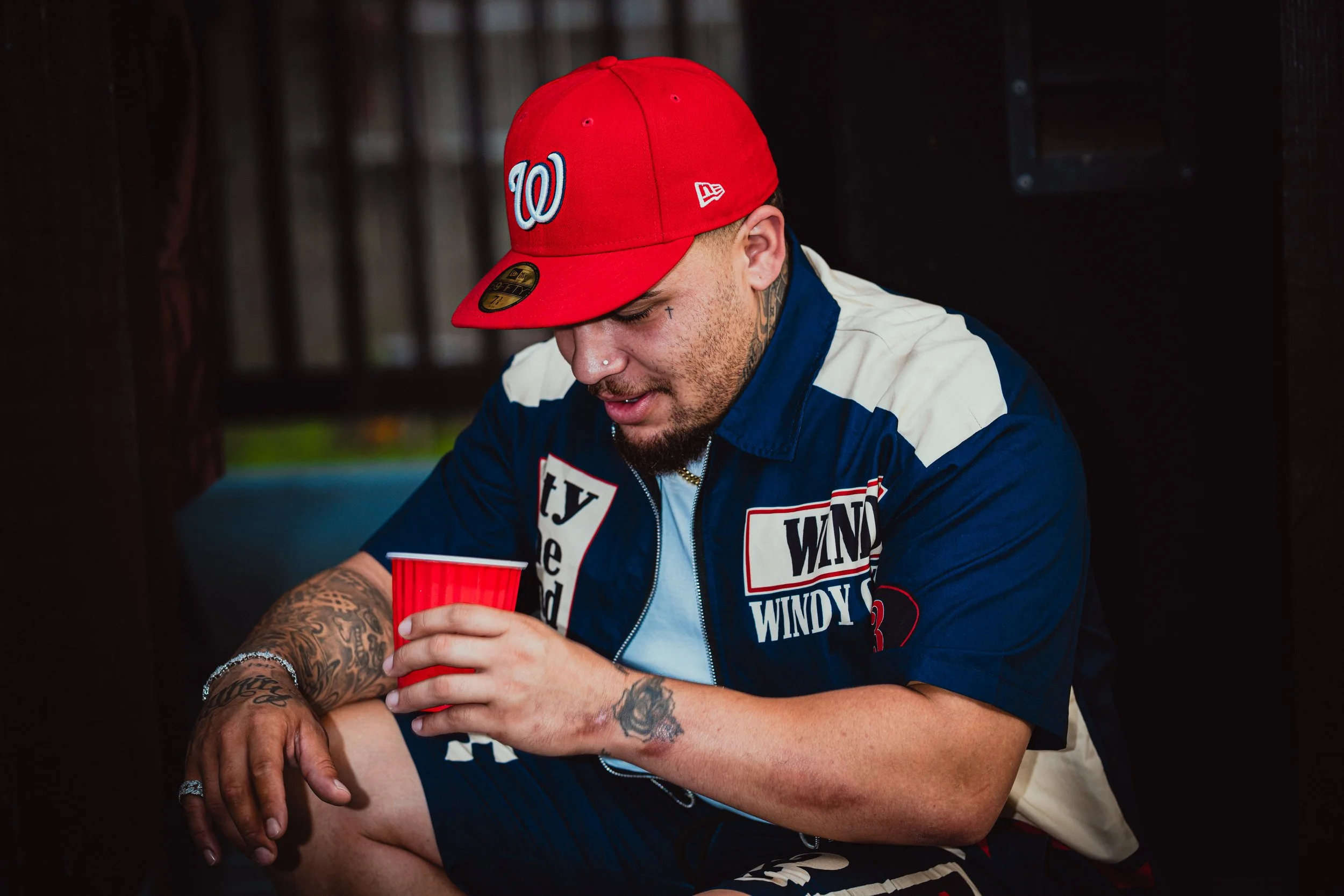 A man wearing a red baseball cap with a Washington Nationals logo, tattoos on his arm, and a navy jacket, is sitting and holding a red plastic cup. He is looking down with a slight smile.
