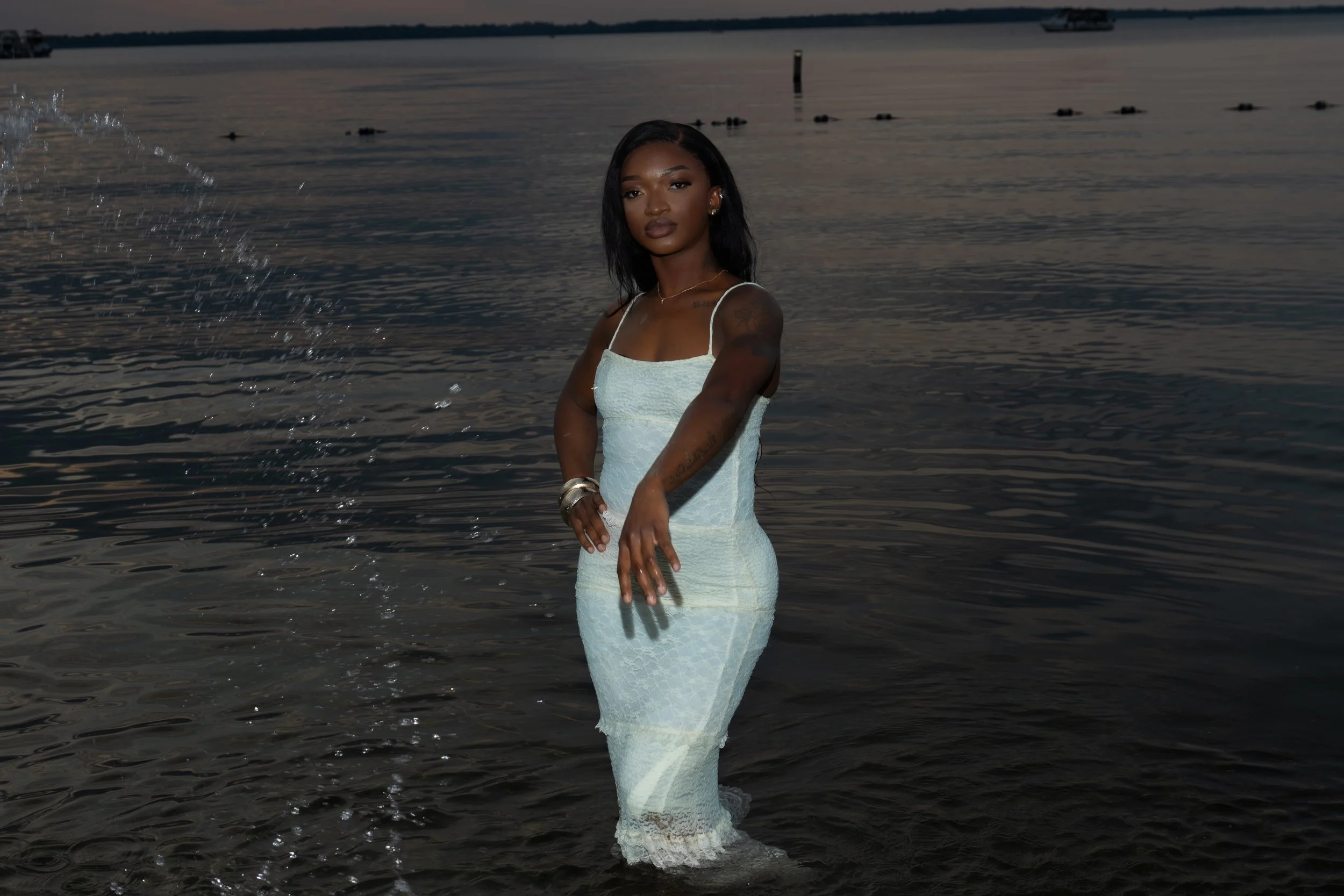 A woman in a white dress standing in water, with a calm body of water and boats in the background at dusk.