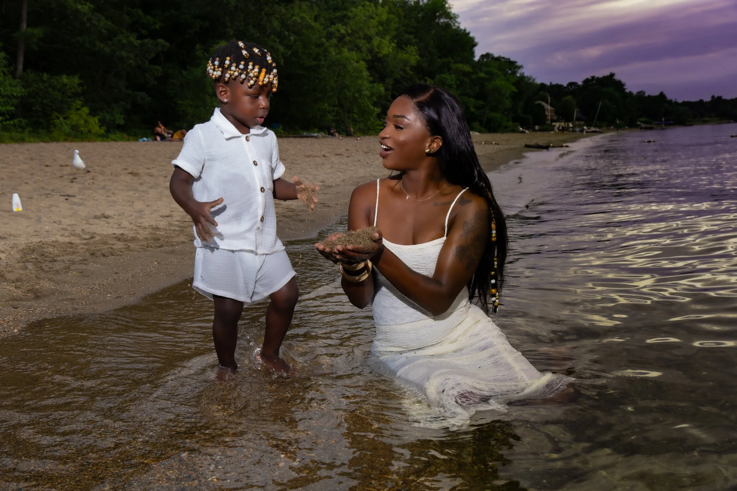 A woman in a white dress kneels in shallow water at the beach, holding sand in her hands, while a young girl in a white outfit with beads in her hair stands nearby, playing with sand. The scene is set at dusk with trees in the background and a cloudy