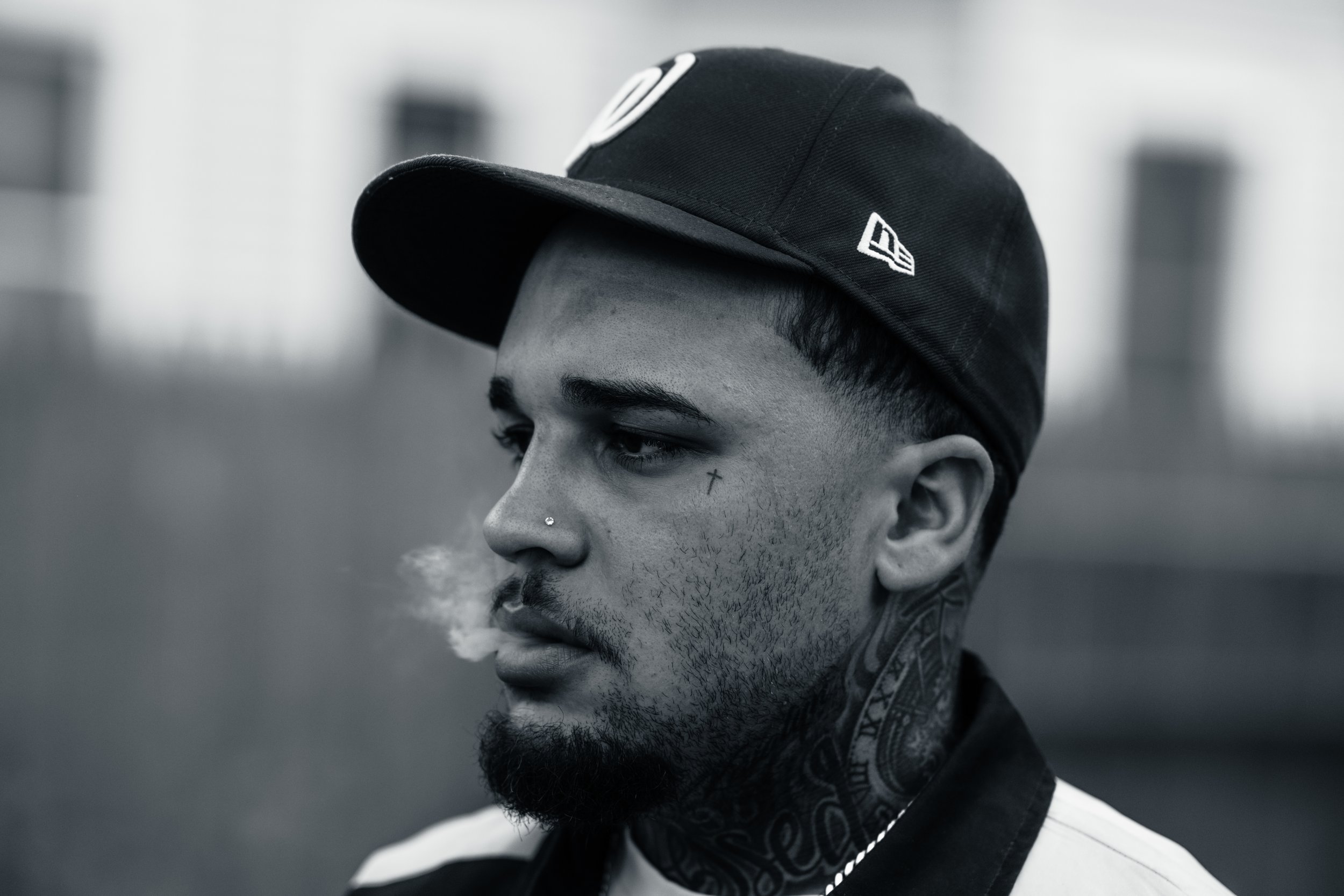 A young man with a beard and tattoos on his neck, wearing a black baseball cap and a scarf with a paisley pattern, is exhaling smoke or vapor. The photo is in black and white.