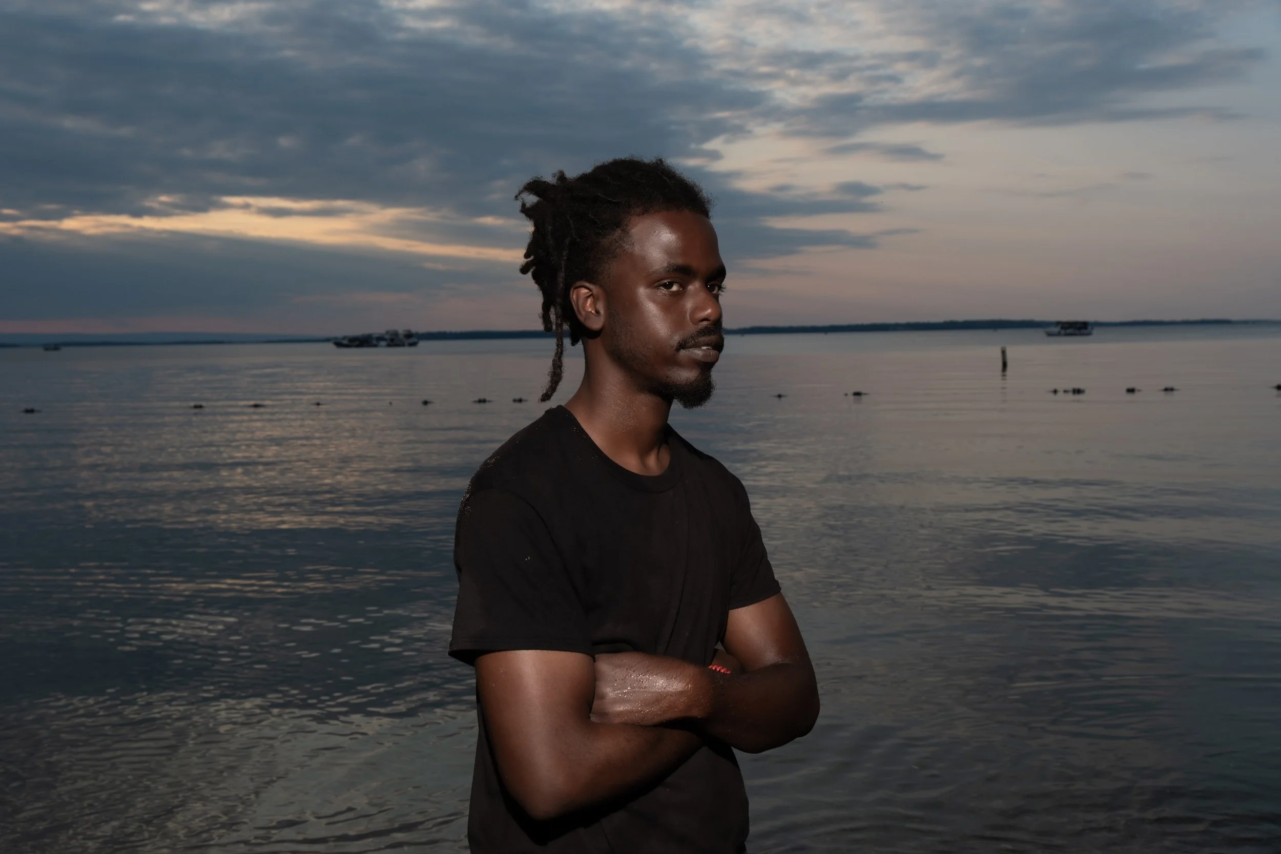 A man with dreadlocks and a beard standing by the water during sunset with a cloudy sky in the background.