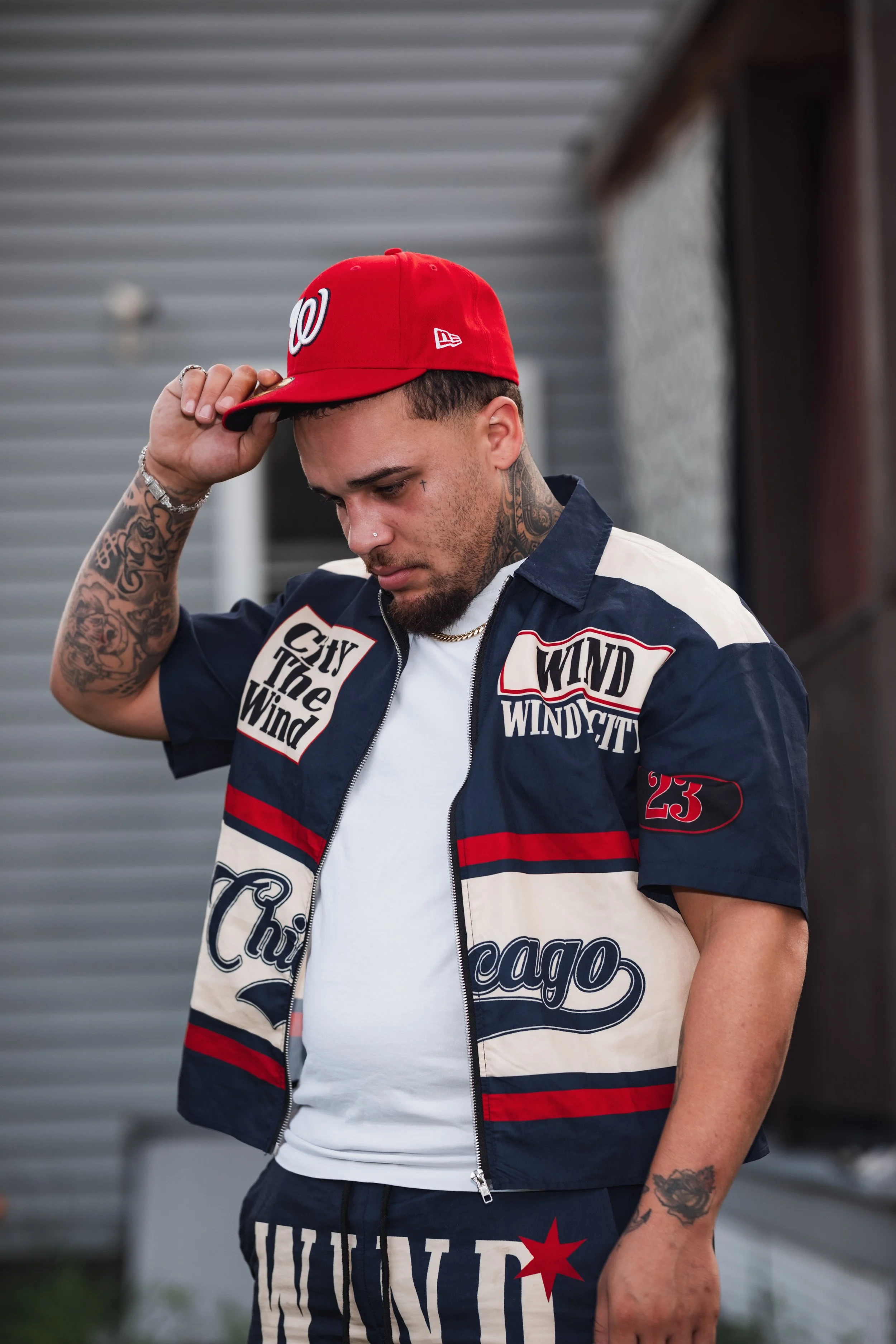 A young man with tattoos on his arm and neck, wearing a red Washington Nationals baseball cap and a navy blue, white, and red Chicago-themed jacket, looking down.