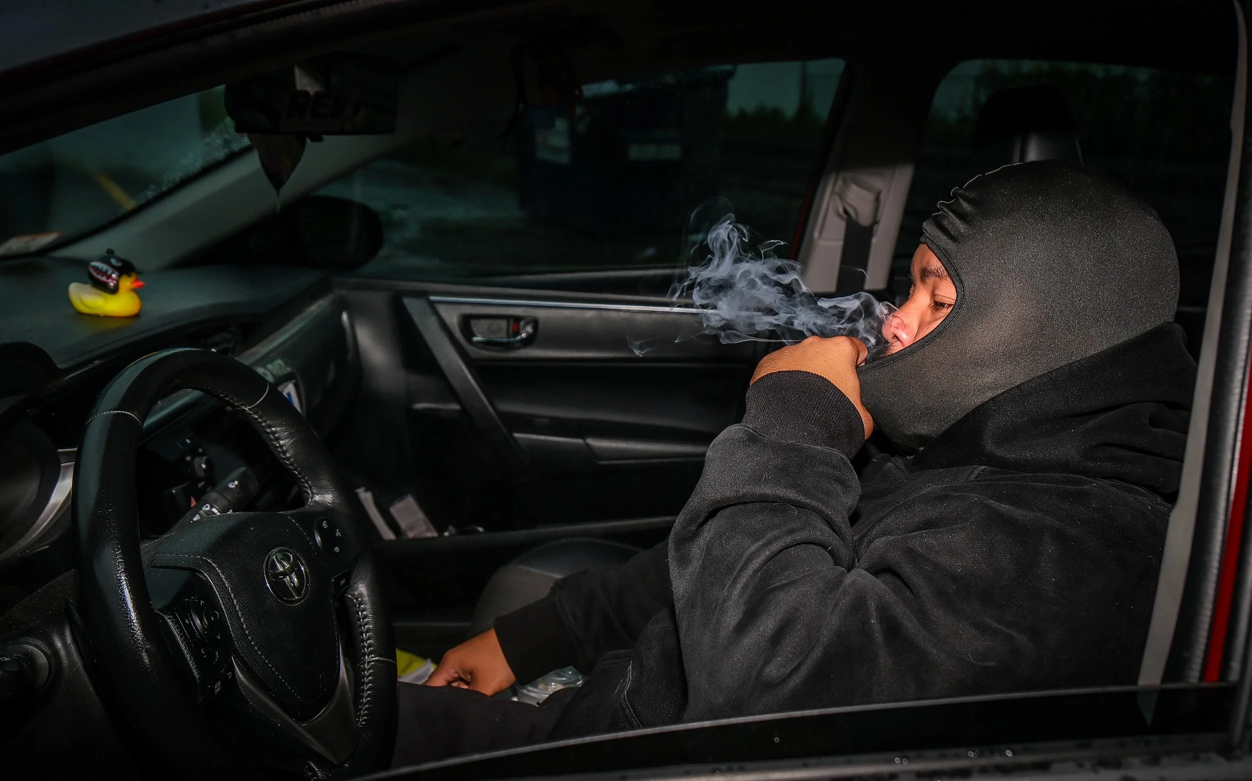 Person wearing a black hoodie and ski mask sitting in the driver seat of a vehicle, exhaling smoke or vapor. The vehicle's interior, including steering wheel and dashboard, is visible.