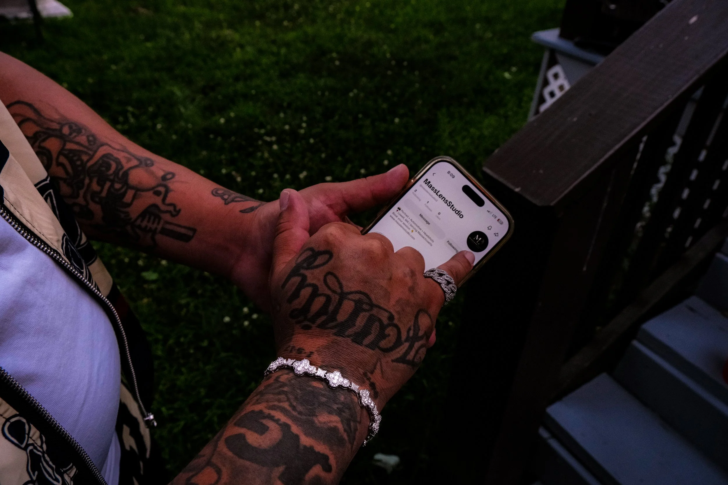 Person with tattoos using a smartphone at an outdoor gathering, with a grassy area and a cooler in the background.