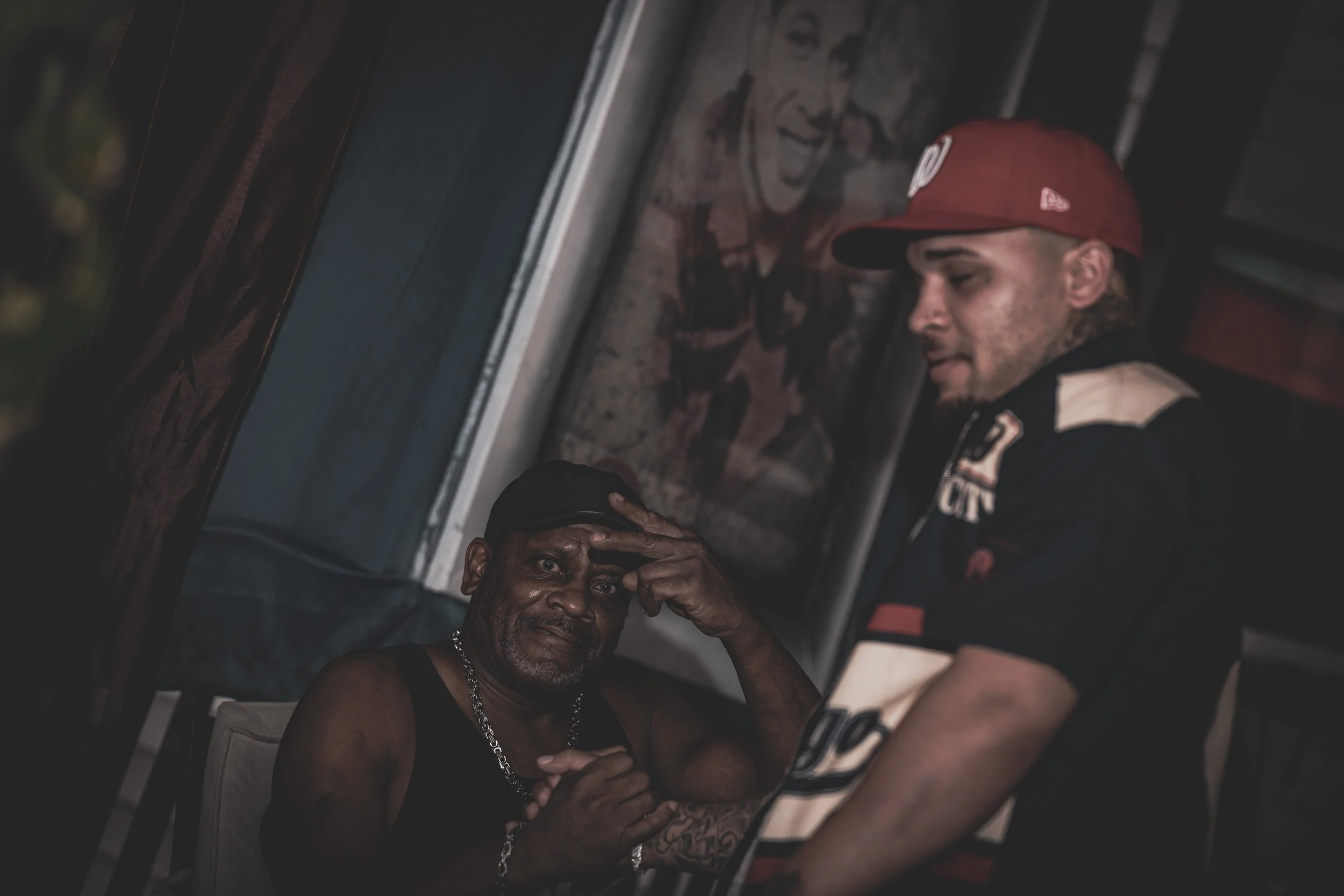 A tattooed man with a chain around his neck sitting on a chair, looking distressed while a person in a red cap and shirt leans over him, holding his head in support.