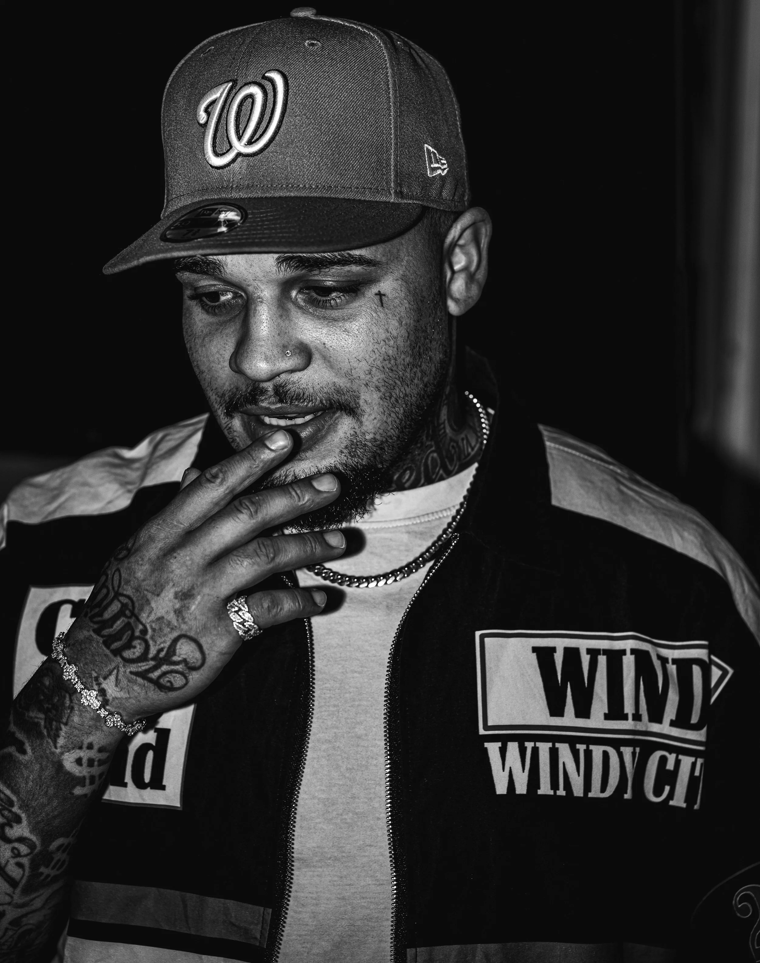 Black and white photo of a man wearing a baseball cap, with tattoos on his hand and face, and a hoodie with text, touching his chin with his fingers.