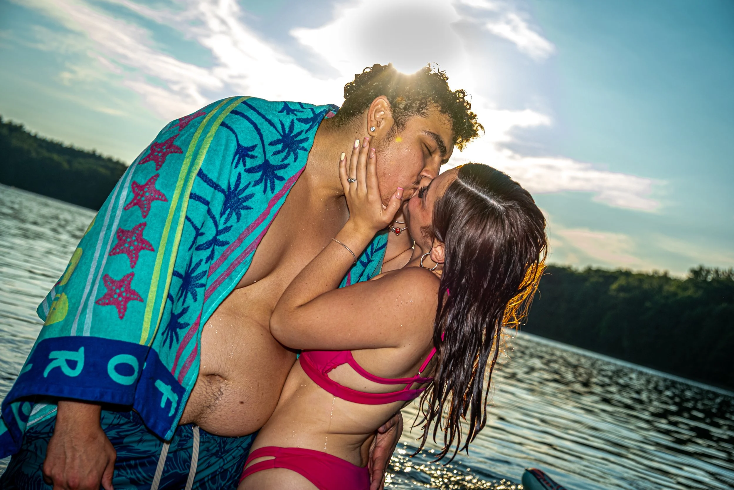 A young couple kissing near a body of water during sunset, with the sun behind the man and a partly cloudy sky.