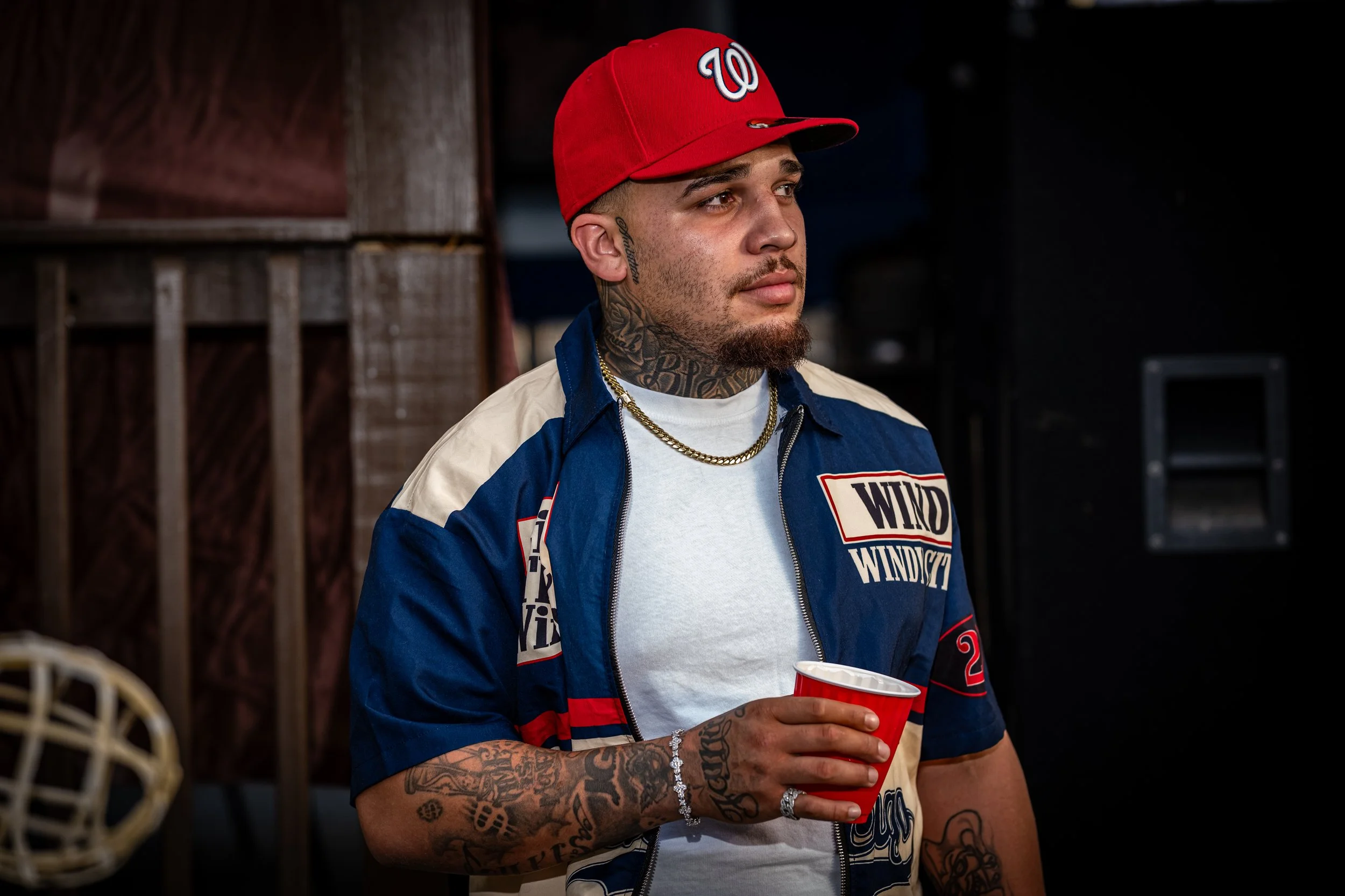 A man with tattoos, wearing a red baseball cap with a white Washington Nationals logo, a navy blue and white jacket, a white T-shirt, and jewelry, holding a red cup, standing indoors with wooden walls and dark background.