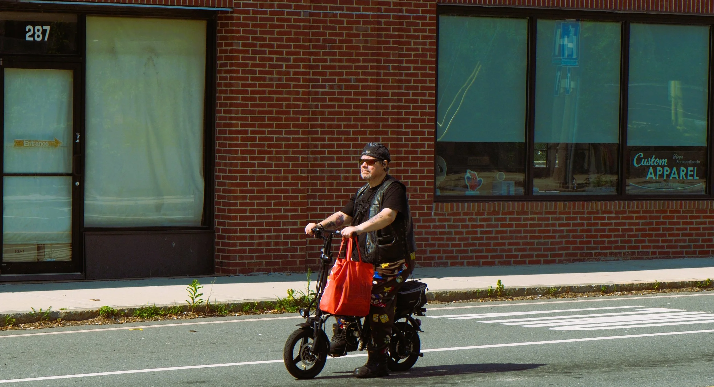 A man riding a bicycle on the street near a brick building with large windows. The man is wearing sunglasses, a black cap, and a black leather vest, with tattoos on his arms. The building has signs for a custom apparel store.