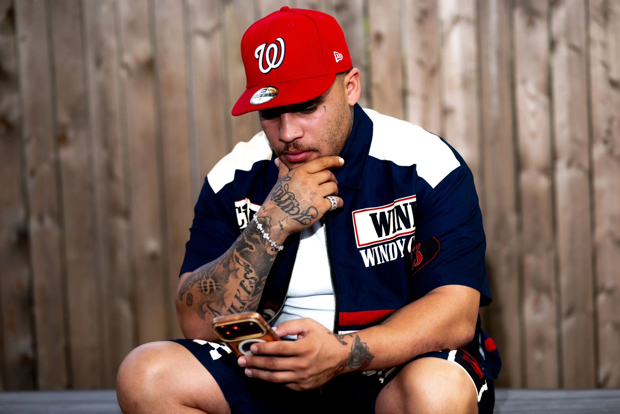 Man wearing a red baseball cap with a white W logo, sitting and looking at his phone, with a wooden fence in the background.