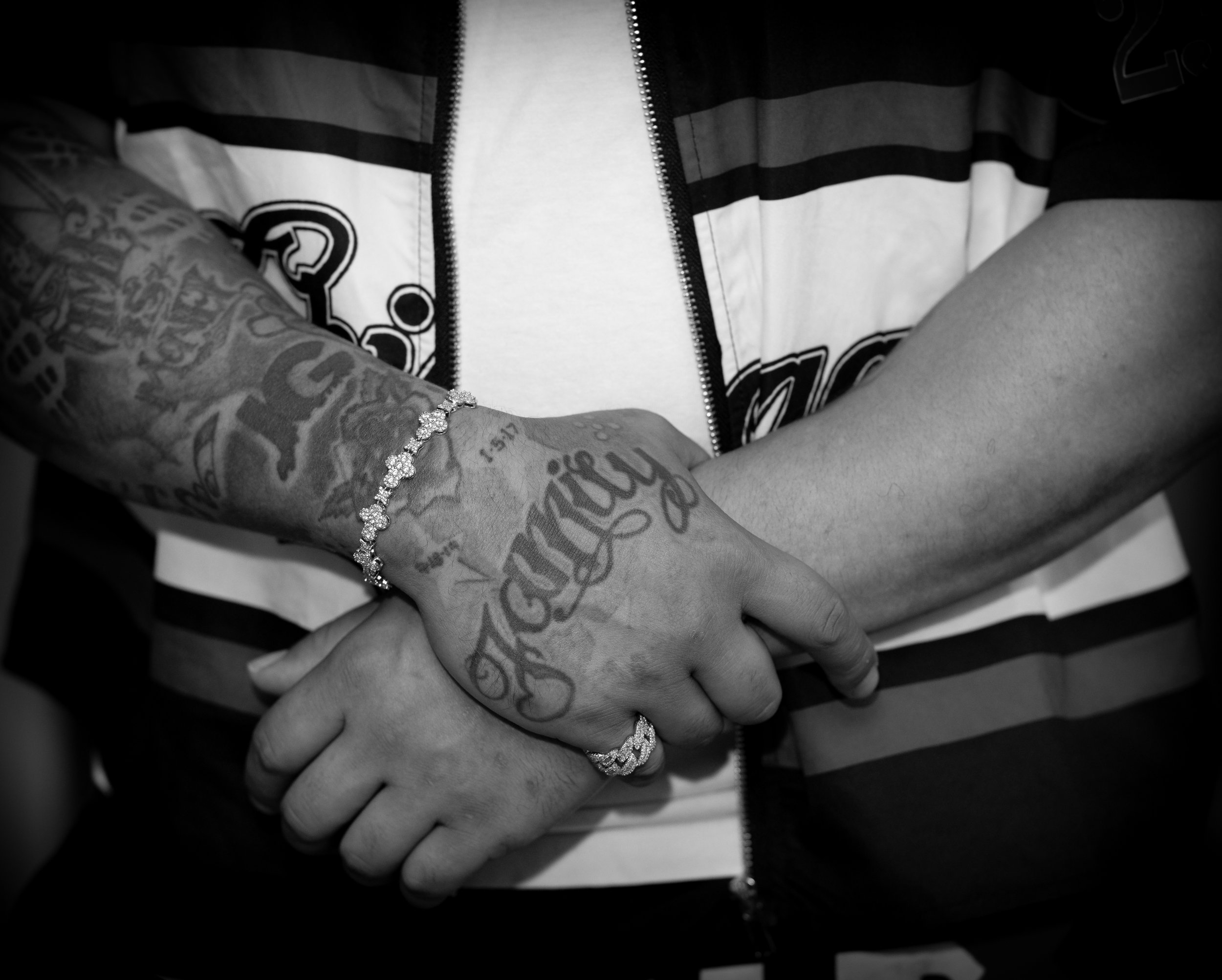 Close-up of two hands clasped together, one with tattoos and jewelry, including rings and a bracelet, and the other hand is bare. The person with tattoos is wearing a zip-up jacket with horizontal stripes.