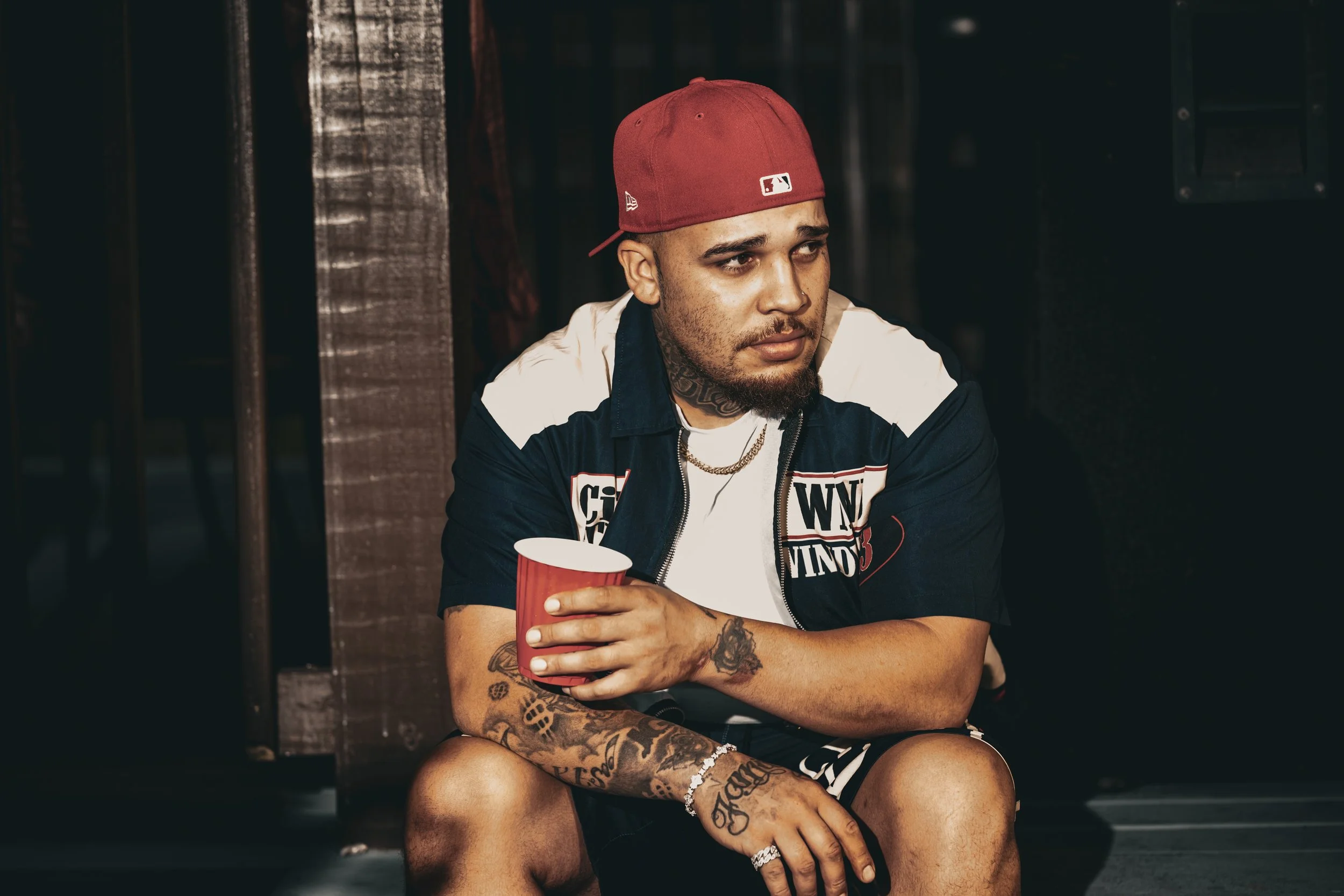 Young man with tattoos wearing a red baseball cap and a NFL jacket, sitting and holding a red cup, in an indoor setting with dark wooden walls.