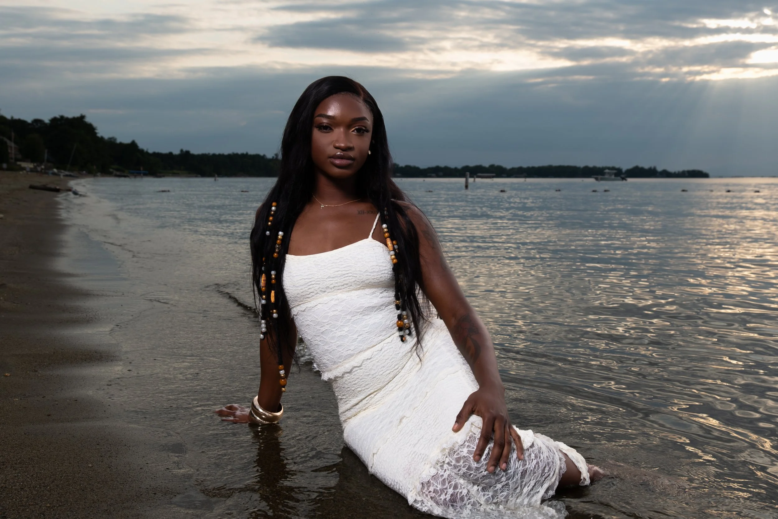 A woman in a white lace dress with beads in her braided hair, sitting on the sandy shore of a body of water during sunset, with a cloudy sky and a boat in the distance.