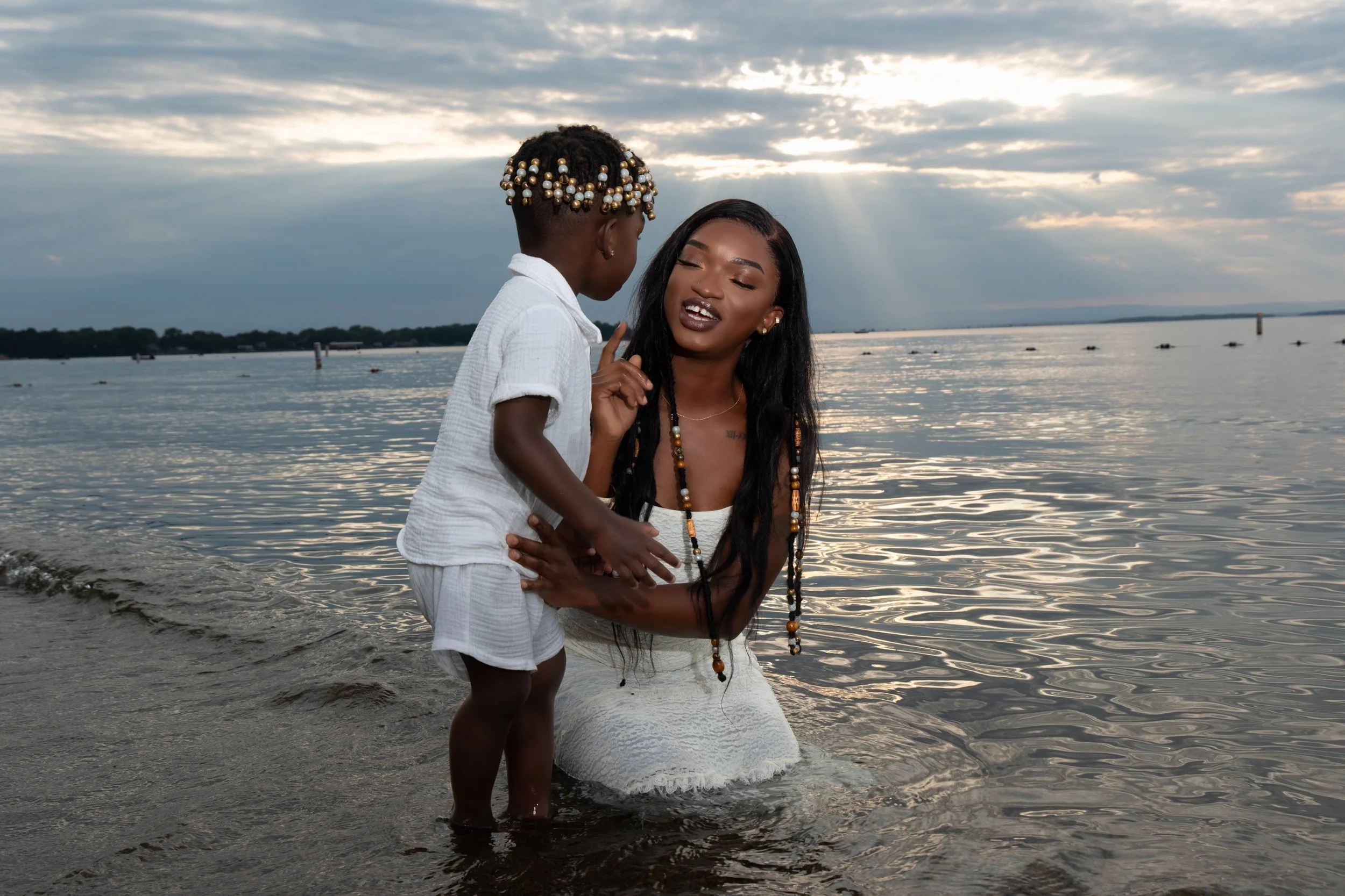Two women embrace at the edge of a body of water, with the sky and clouds overhead during sunset or sunrise.
