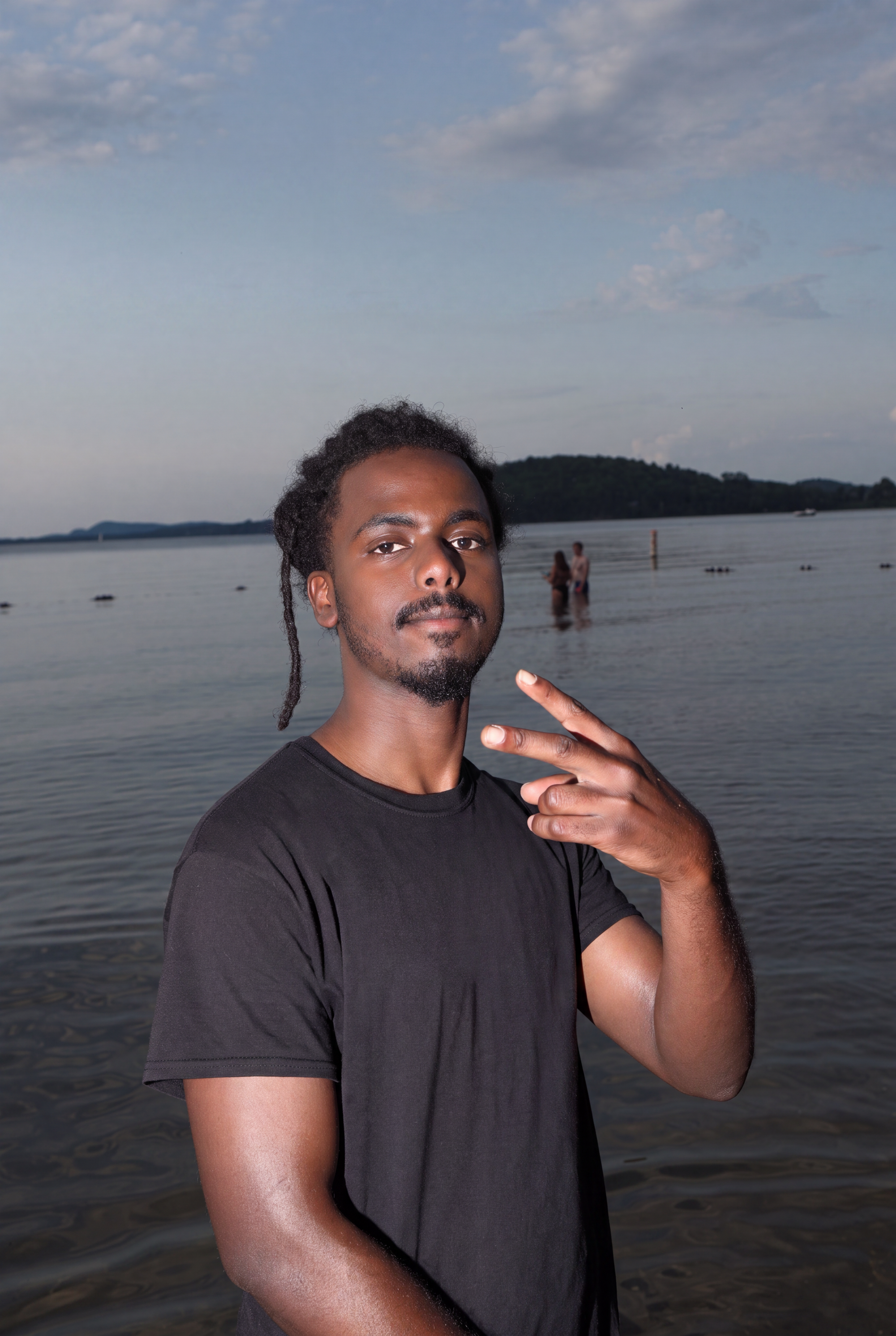 A man with dreadlocks and a mustache posing near a body of water during daytime, with a cloudy sky and hills in the background.