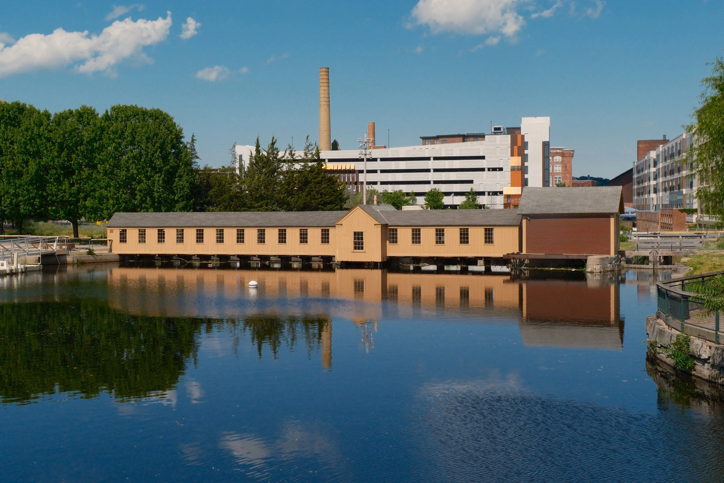 A calm river reflecting buildings and green trees along its banks on a clear, sunny day with a blue sky and scattered clouds.