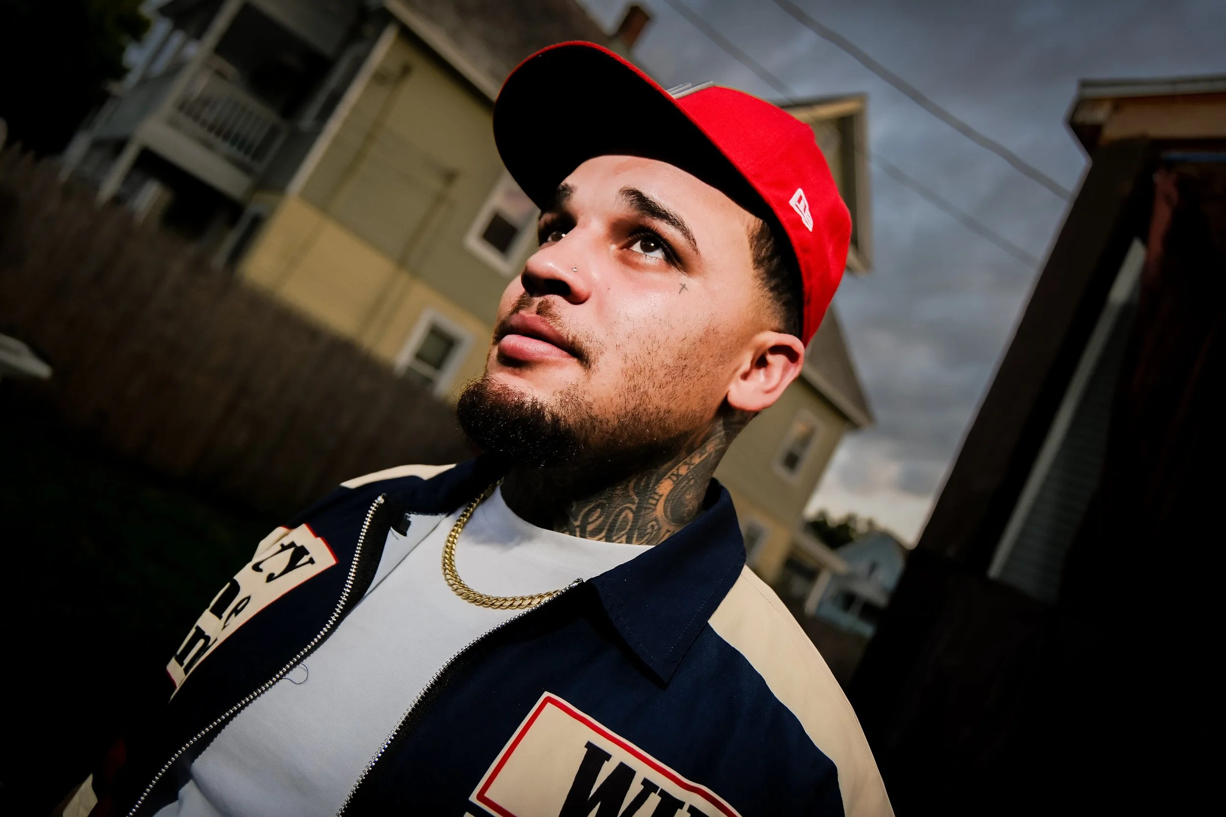 Close-up of a young man wearing a red cap backwards and a jacket with patches, looking slightly upward with a serious expression, in an outdoor urban setting during dusk or early evening.
