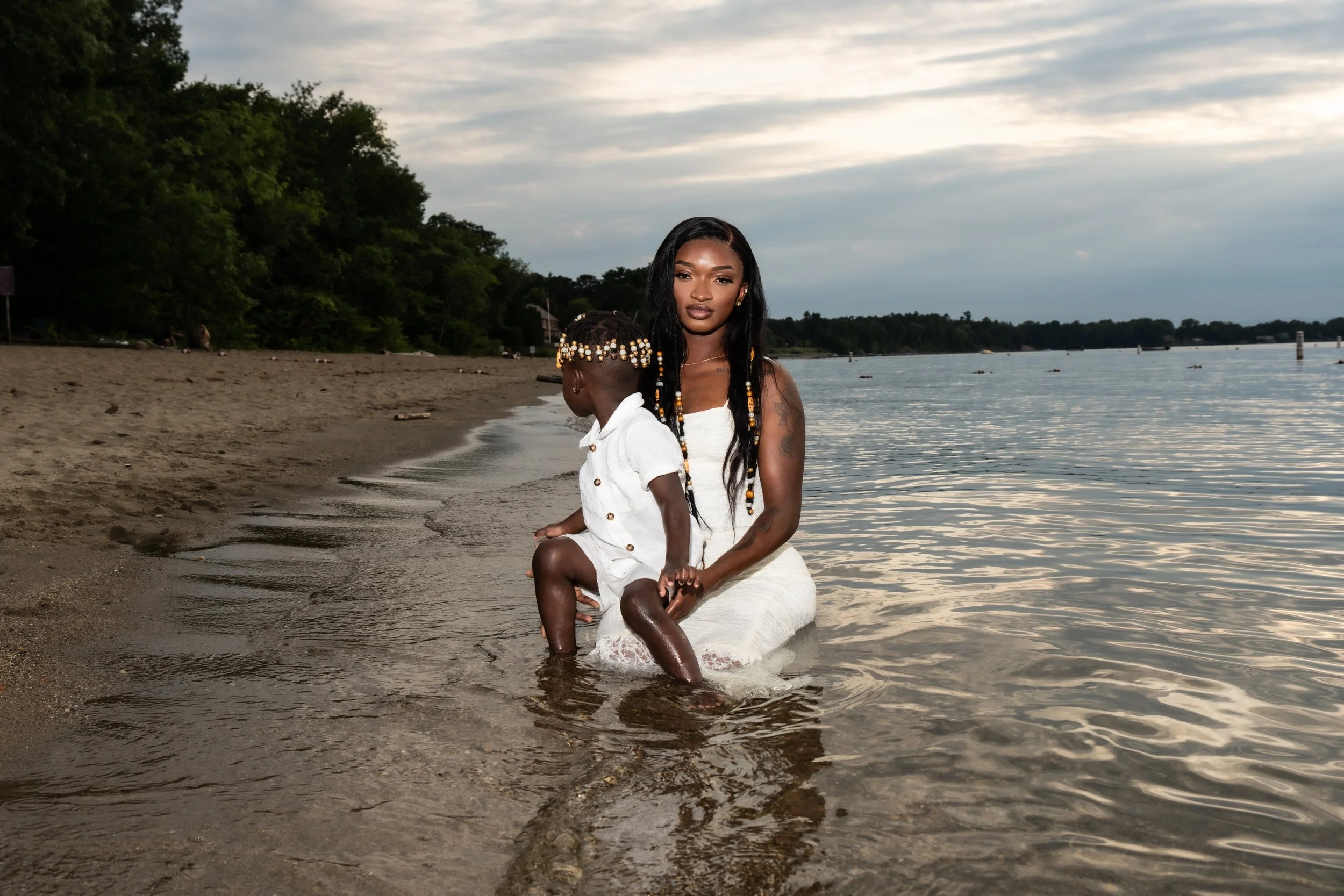 An African American woman with long, black hair and tattoos on her arm, wearing a white dress, is sitting on a white blanket with her young daughter at the edge of a river or lake. The daughter, wearing a white outfit with beads in her hair, is sitti