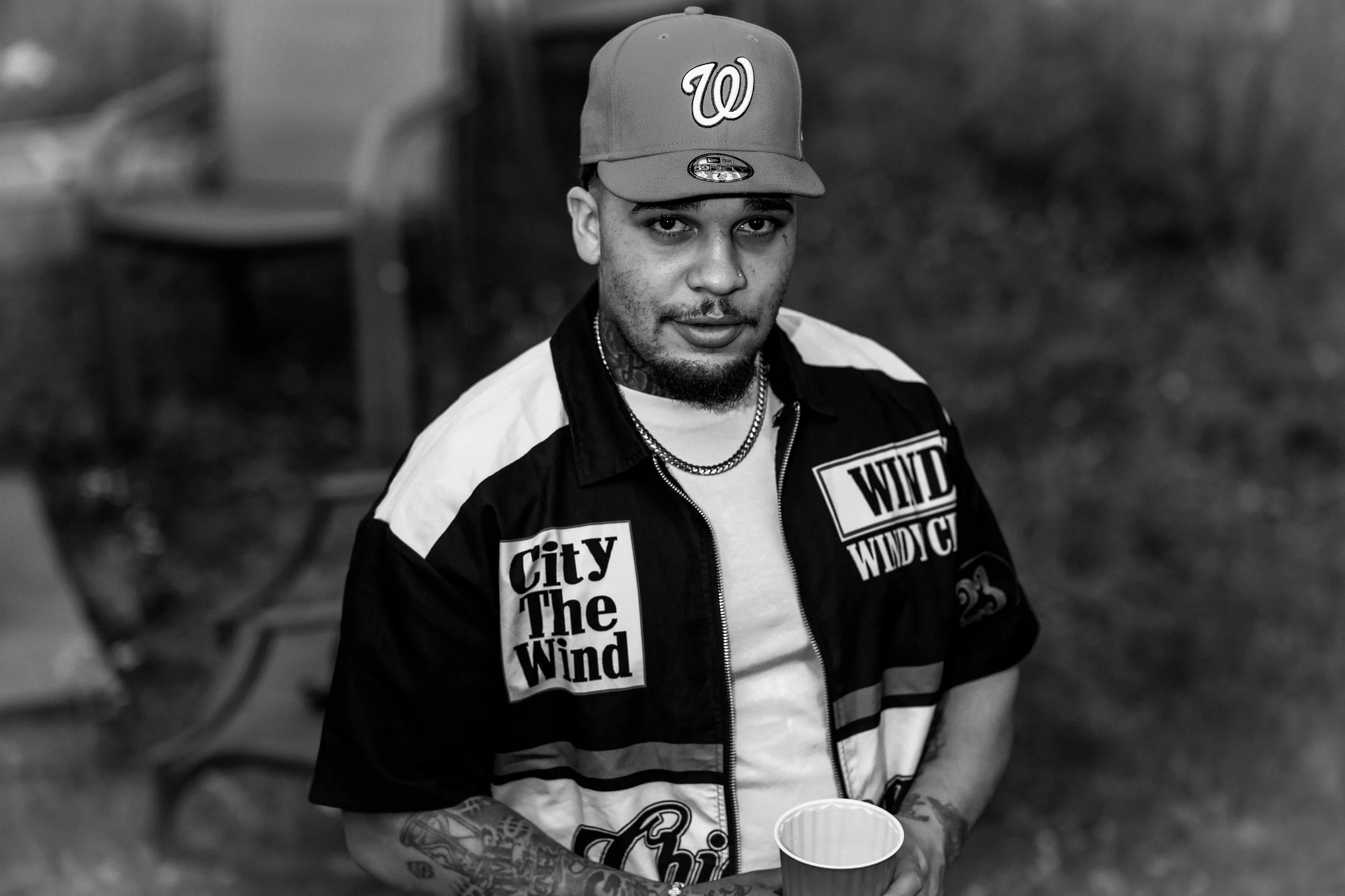 A young man with tattoos on his arm, wearing a baseball cap with a 'W' logo, a Nike windbreaker with bold text, and chains around his neck, standing outdoors with blurred background.