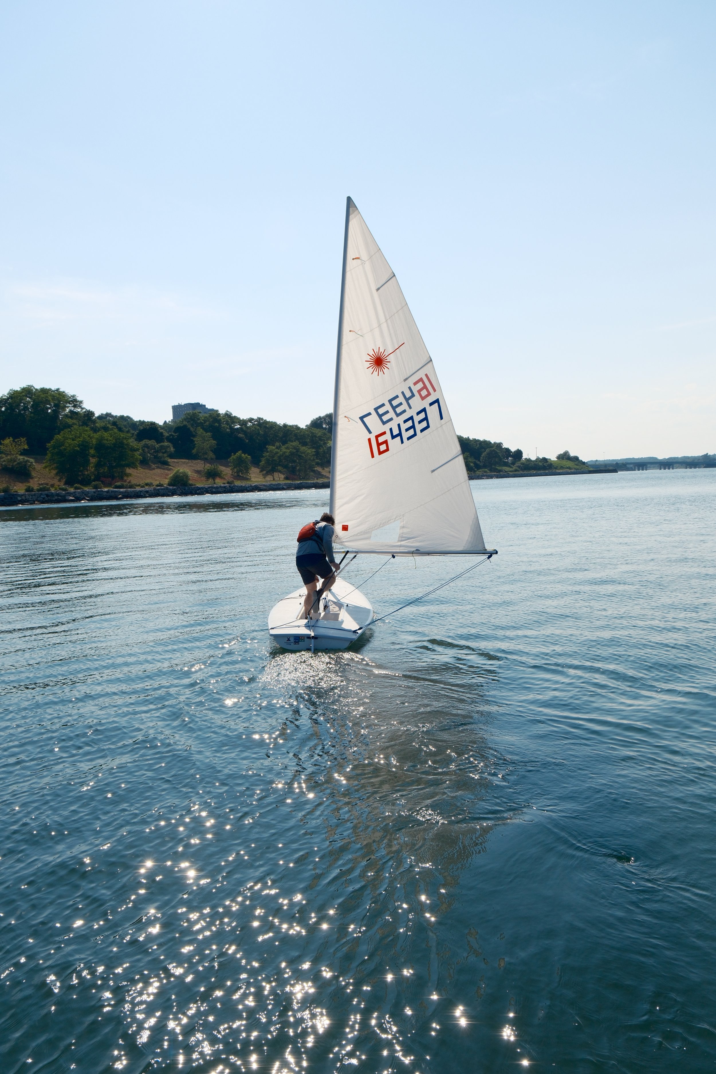A person sailing a small sailboat on a calm body of water during daytime with tree-covered hills and a distant bridge in the background.