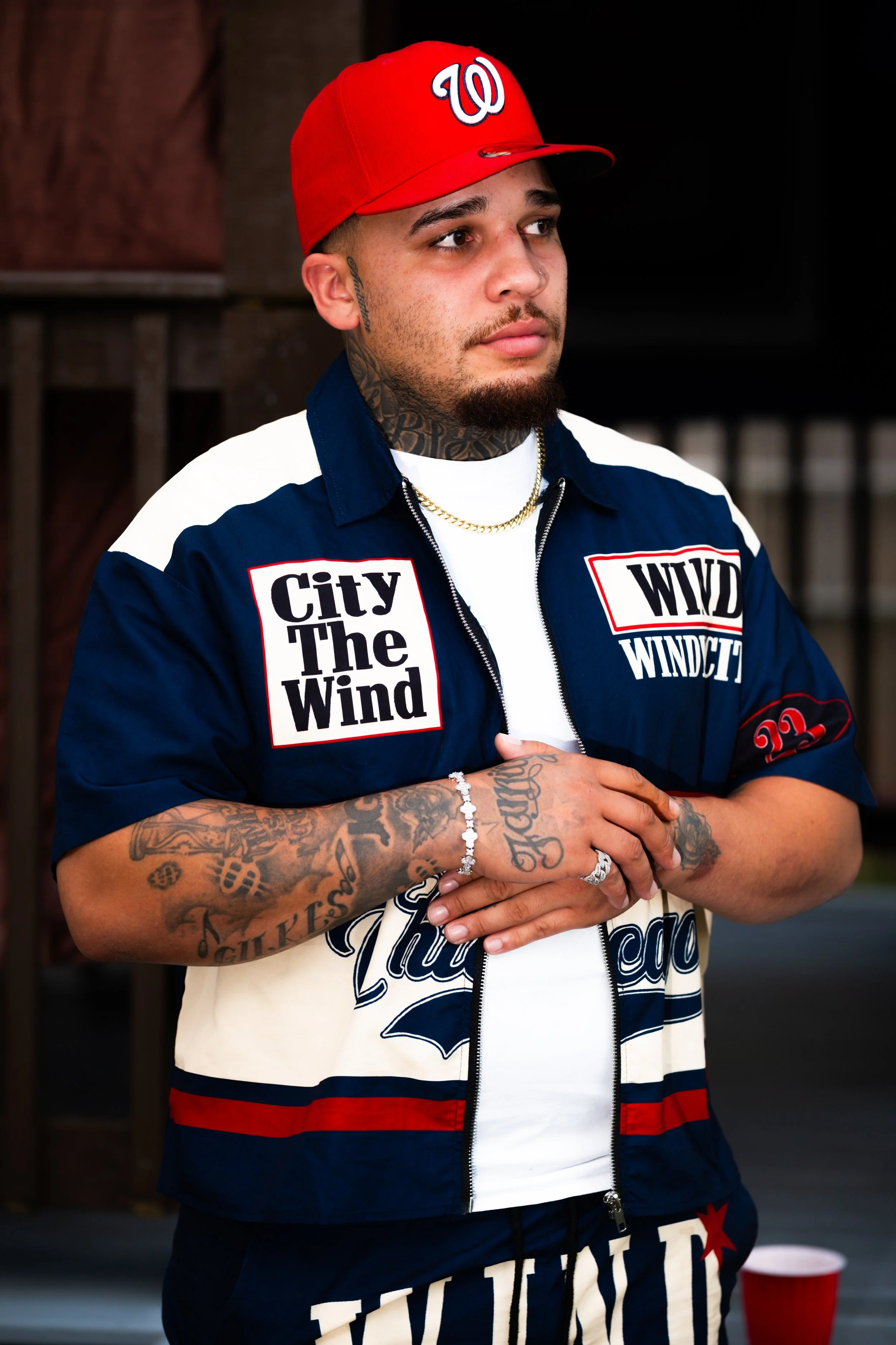 Young man with tattoos wearing a red Washington Nationals cap, a blue and white windbreaker with patches, a white T-shirt, and gold chains, standing outdoors with a neutral expression.