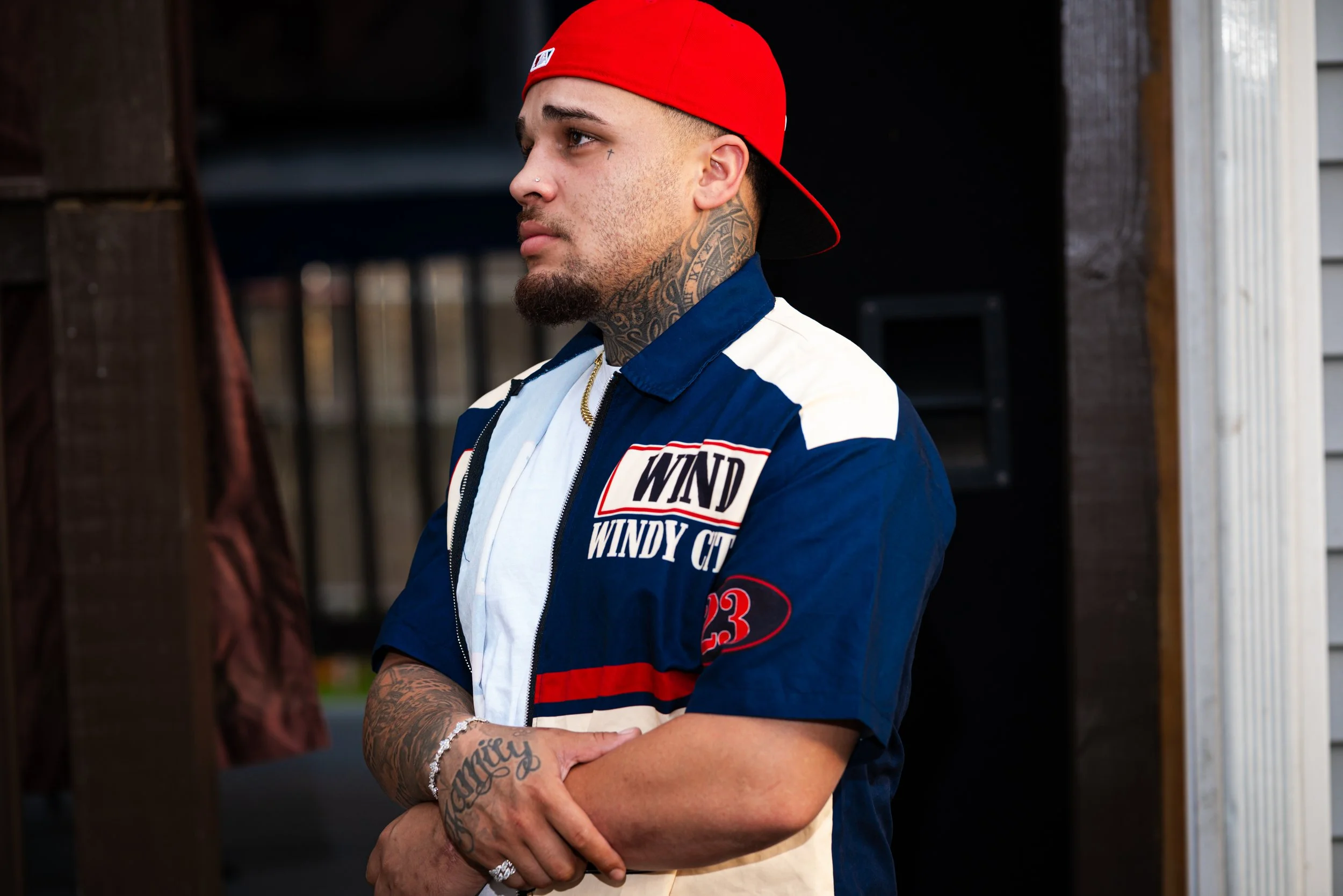 Young man with tattoos on his neck and hands wearing a red cap backwards and a blue and white jacket with patches, standing with arms crossed in an outdoor setting.
