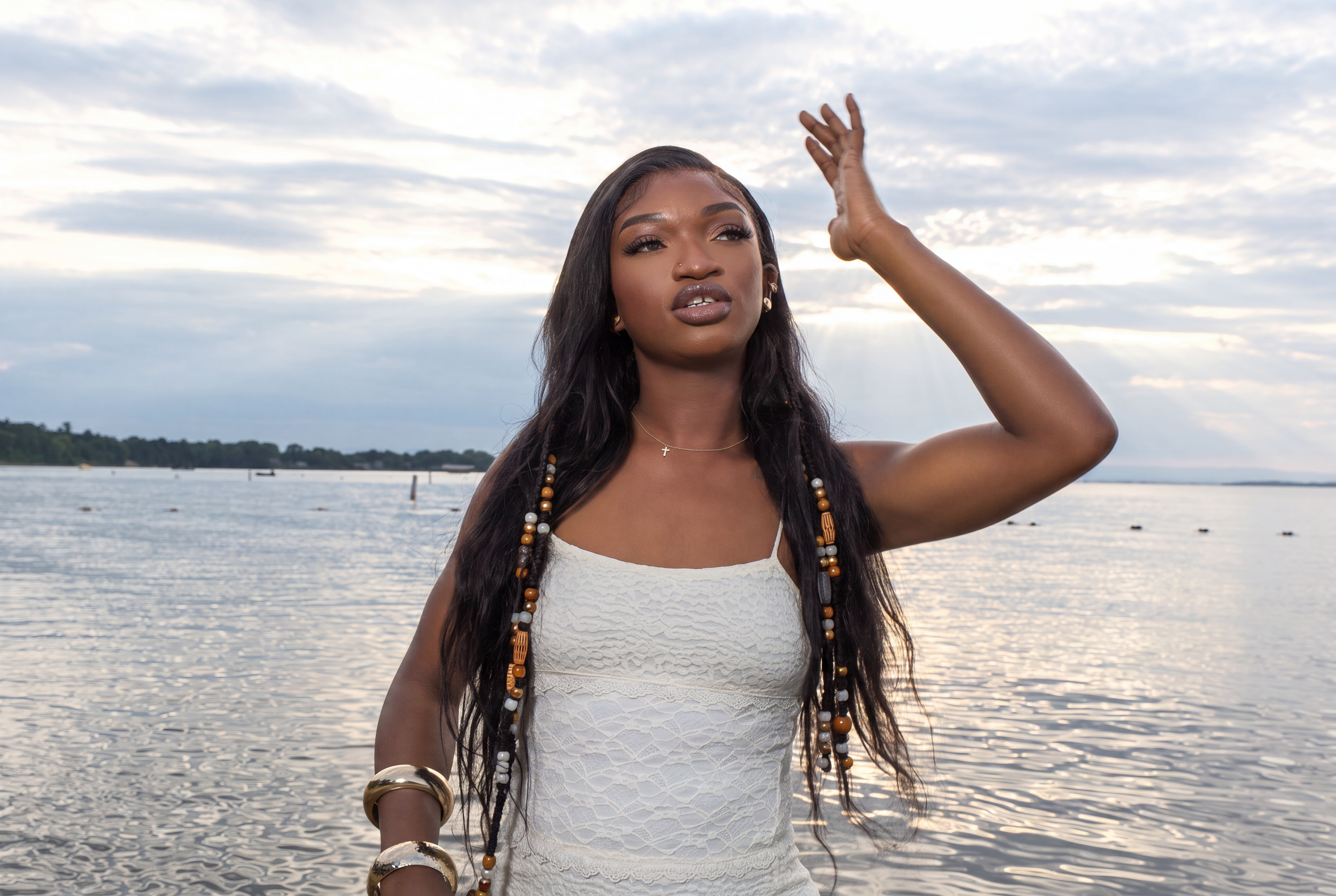 A woman with long dark hair and jewelry standing near a body of water during sunset or sunrise.