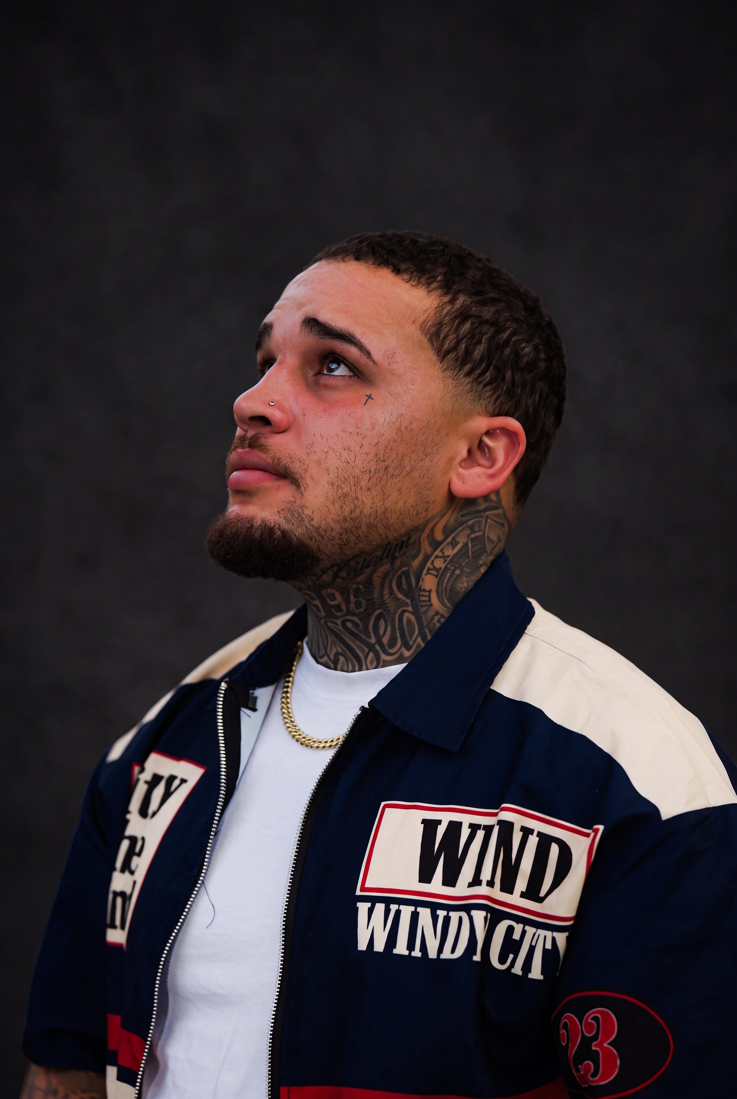 A young man with short dark hair and tattoos on his neck and face, looking upward. He has a nose piercing and wears a white t-shirt, a gold chain, and a navy blue jacket with patches and bold text.