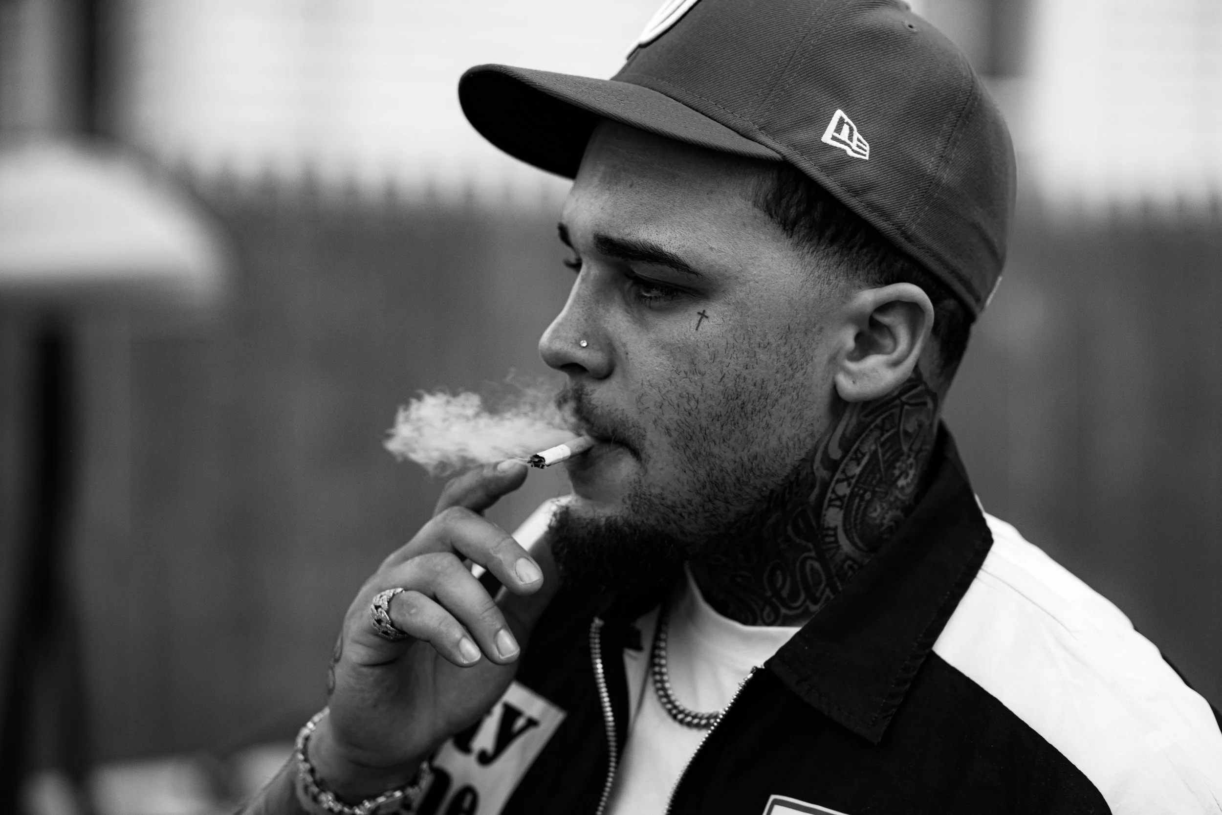A young man with tattoos, piercings, and jewelry, wearing a baseball cap and a zip-up jacket, is smoking a cigarette.