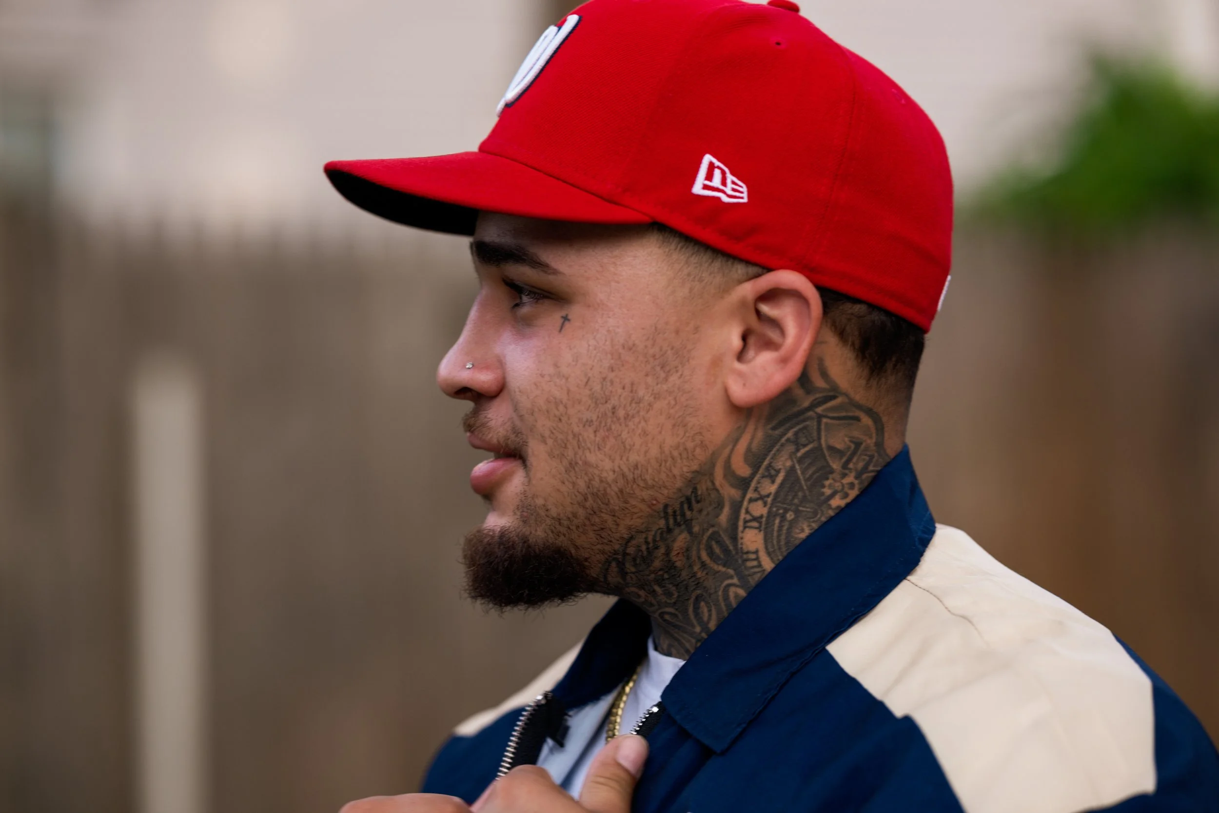 Profile of a young man wearing a red cap with a black underbill, a navy and white jacket, and tattoos on his neck and face.